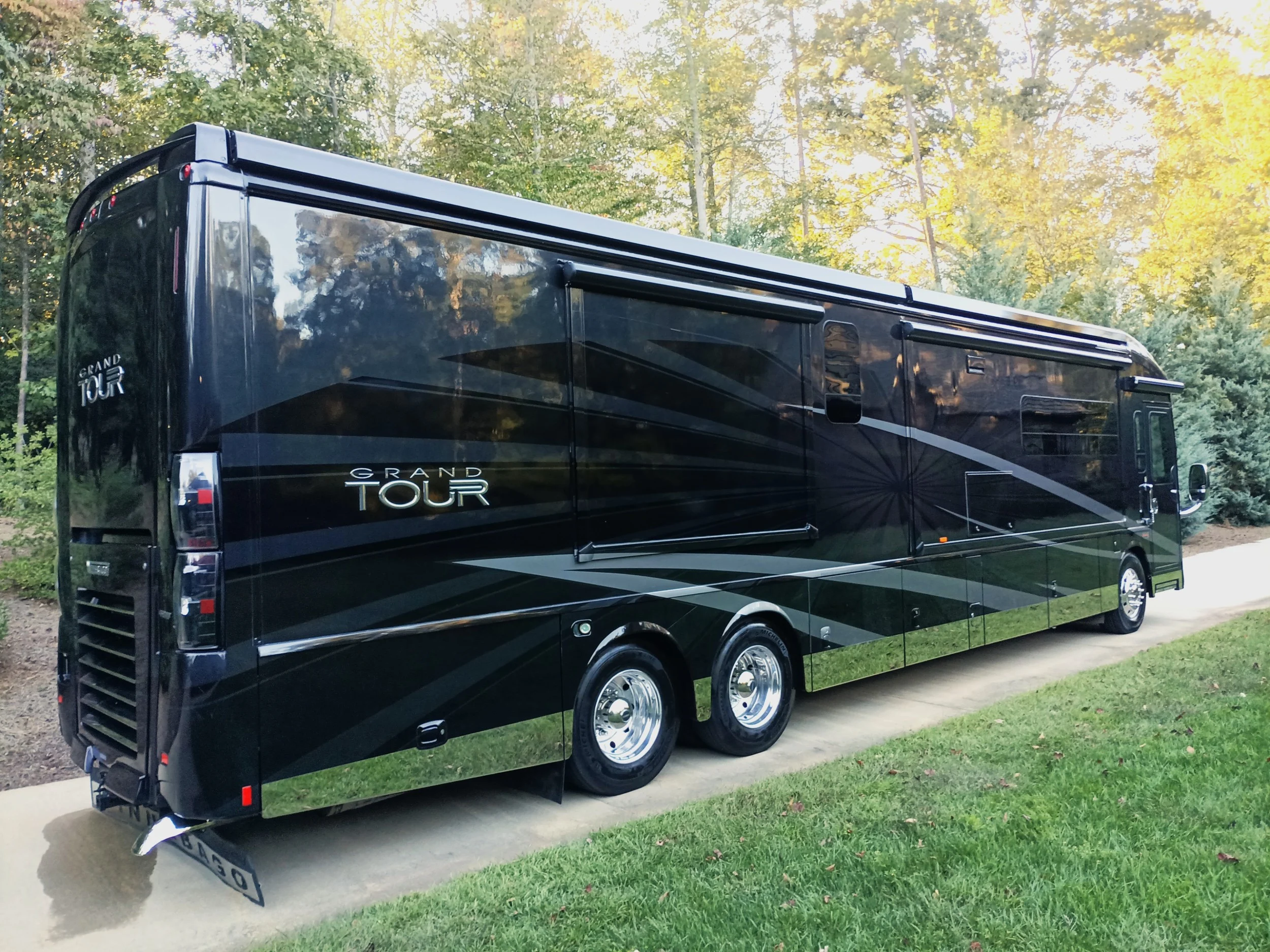 Sleek black motorcoach parked on a driveway, surrounded by lush greenery and trees, with "Grand Tour" branding visible.