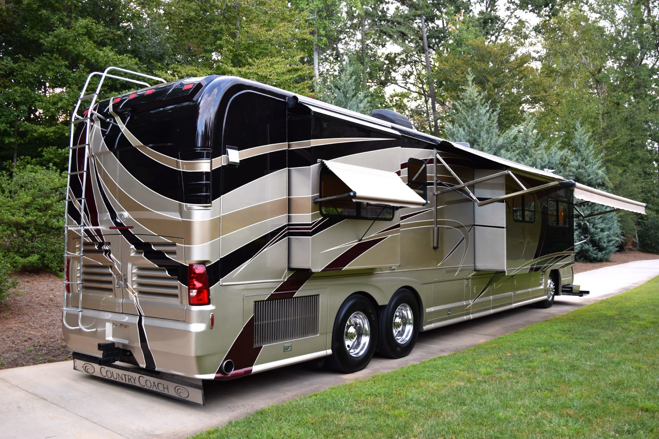 Luxury motorcoach with extended side awning, parked on a paved path in a scenic wooded area.