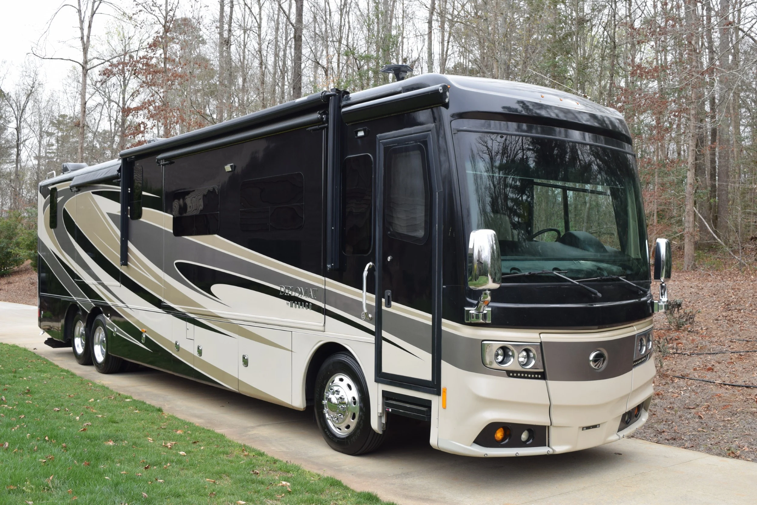 Luxurious Motorcoach and Rvs parked on a driveway near a forest, featuring sleek design and black-beige color scheme.