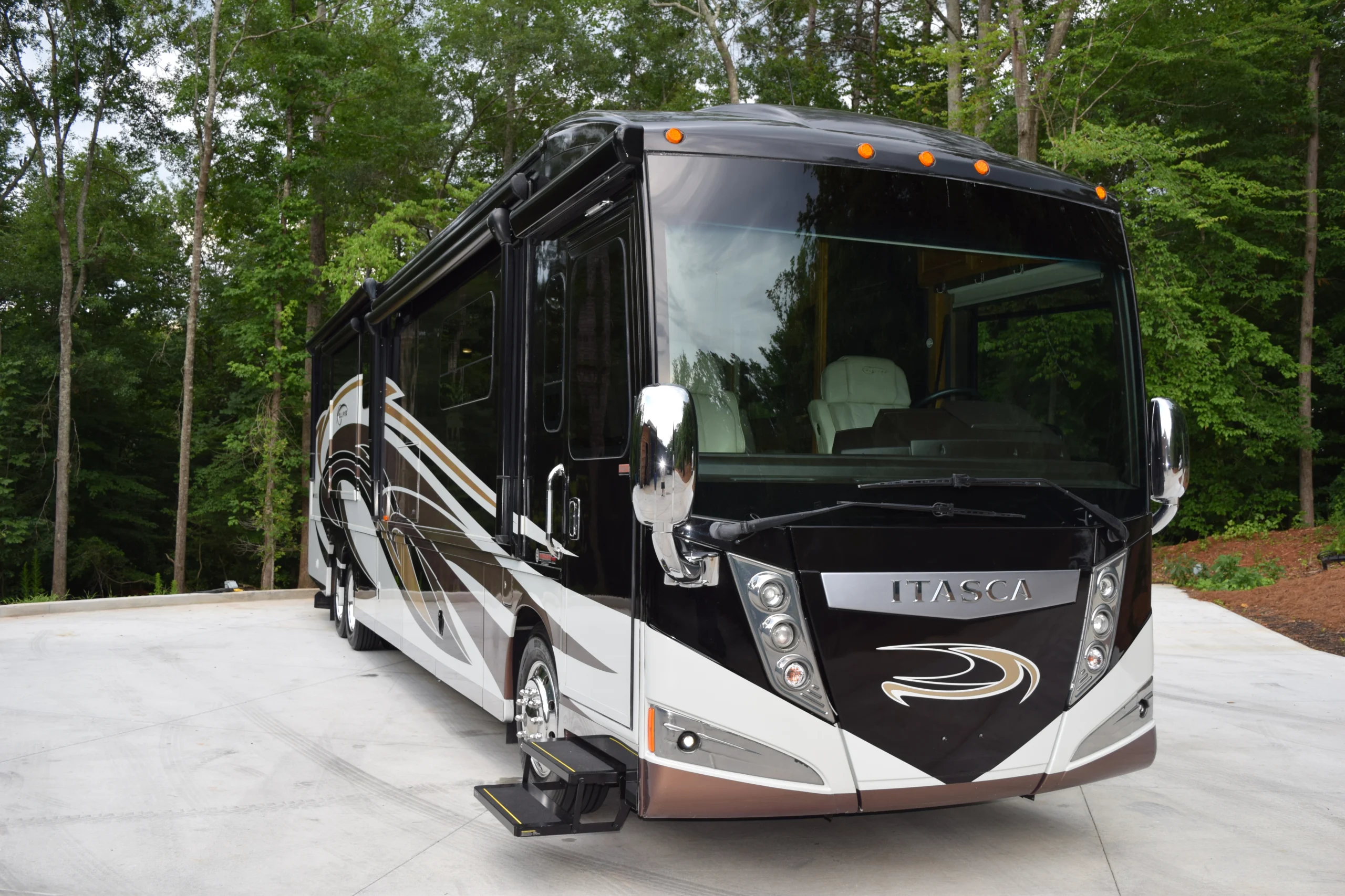 DSC_0524 Luxury motorcoach parked on a concrete driveway, surrounded by lush green trees, showcasing sleek design and reflective surfaces.