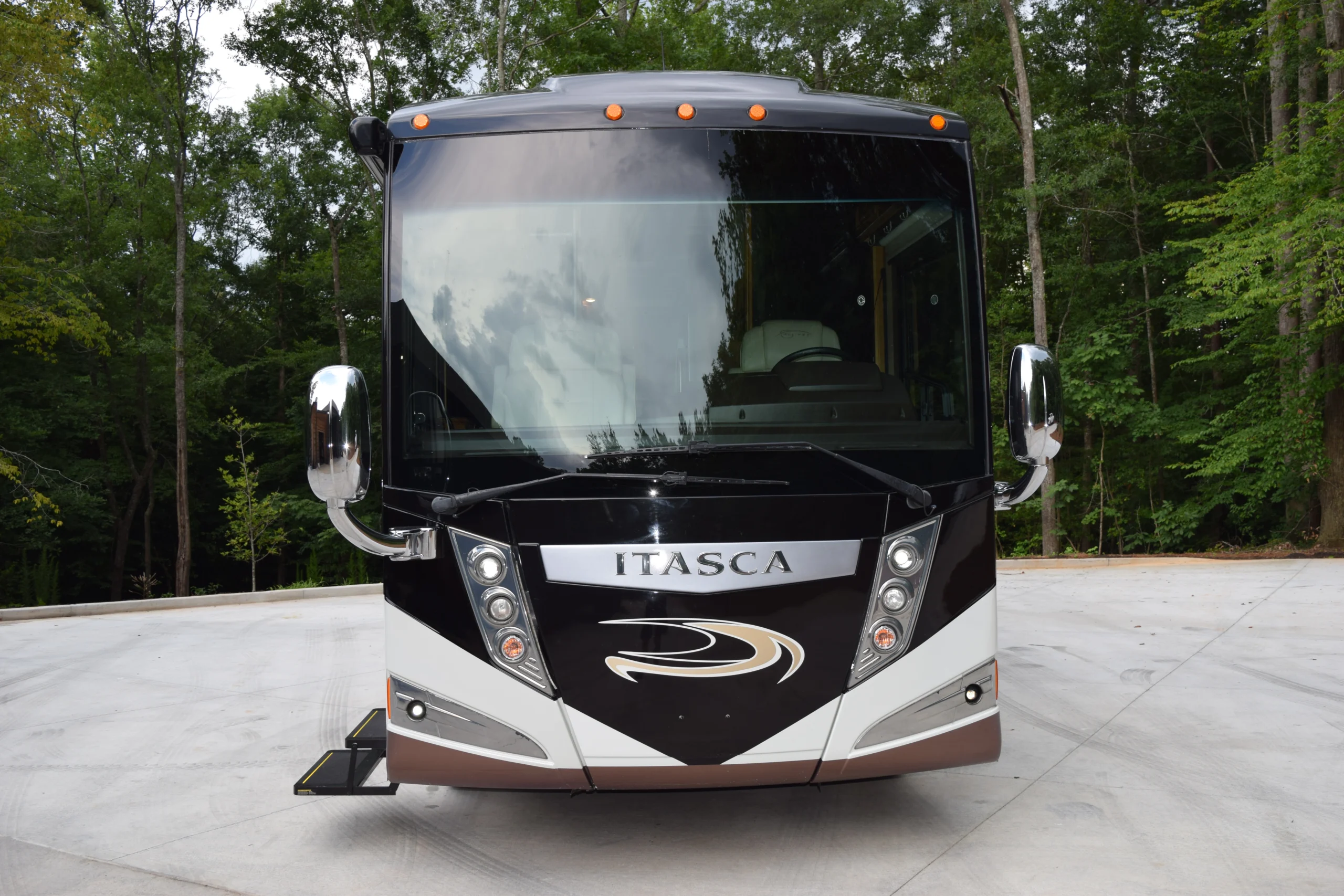 DSC_0526 Front view of a sleek black Itasca motorcoach parked on a paved area, surrounded by lush green trees.