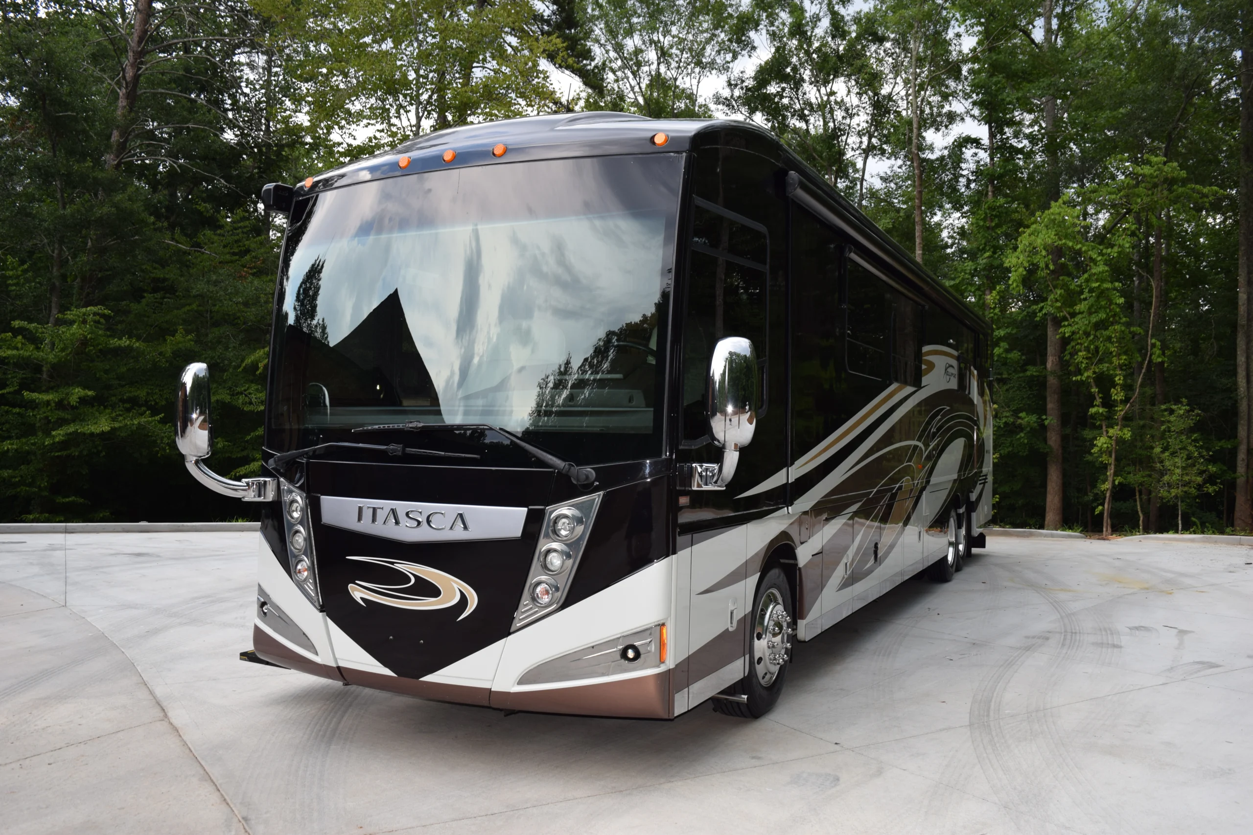 DSC_0527 Black and white motorcoach parked on a concrete driveway surrounded by tall trees, showcasing sleek design and reflective windows.
