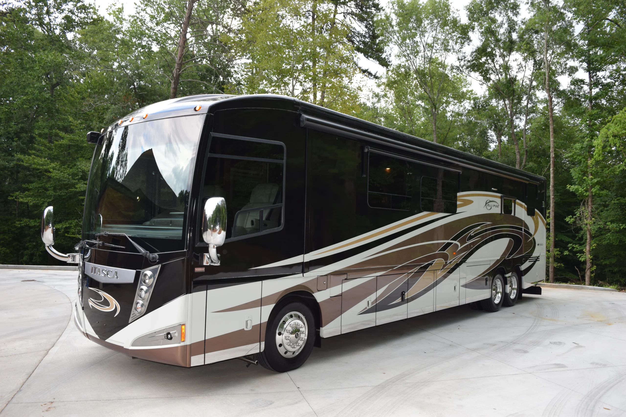 DSC_0528 Luxurious Motorcoach parked on a concrete driveway, surrounded by lush green trees. Brown and white abstract design on side.