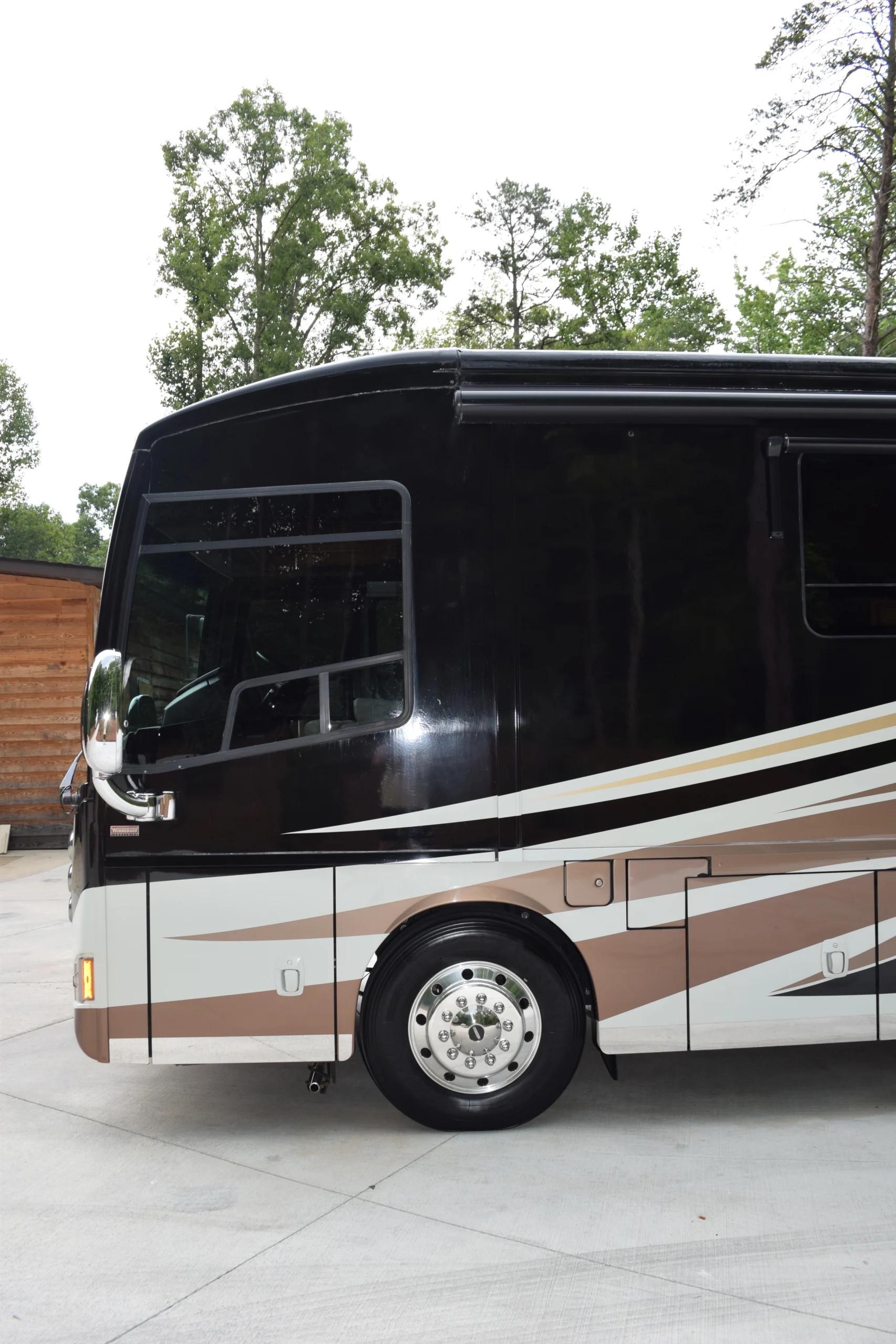 DSC_0529 Rear section of a parked Motorcoach with a glossy black and white design, set against a backdrop of trees and wooden structure.