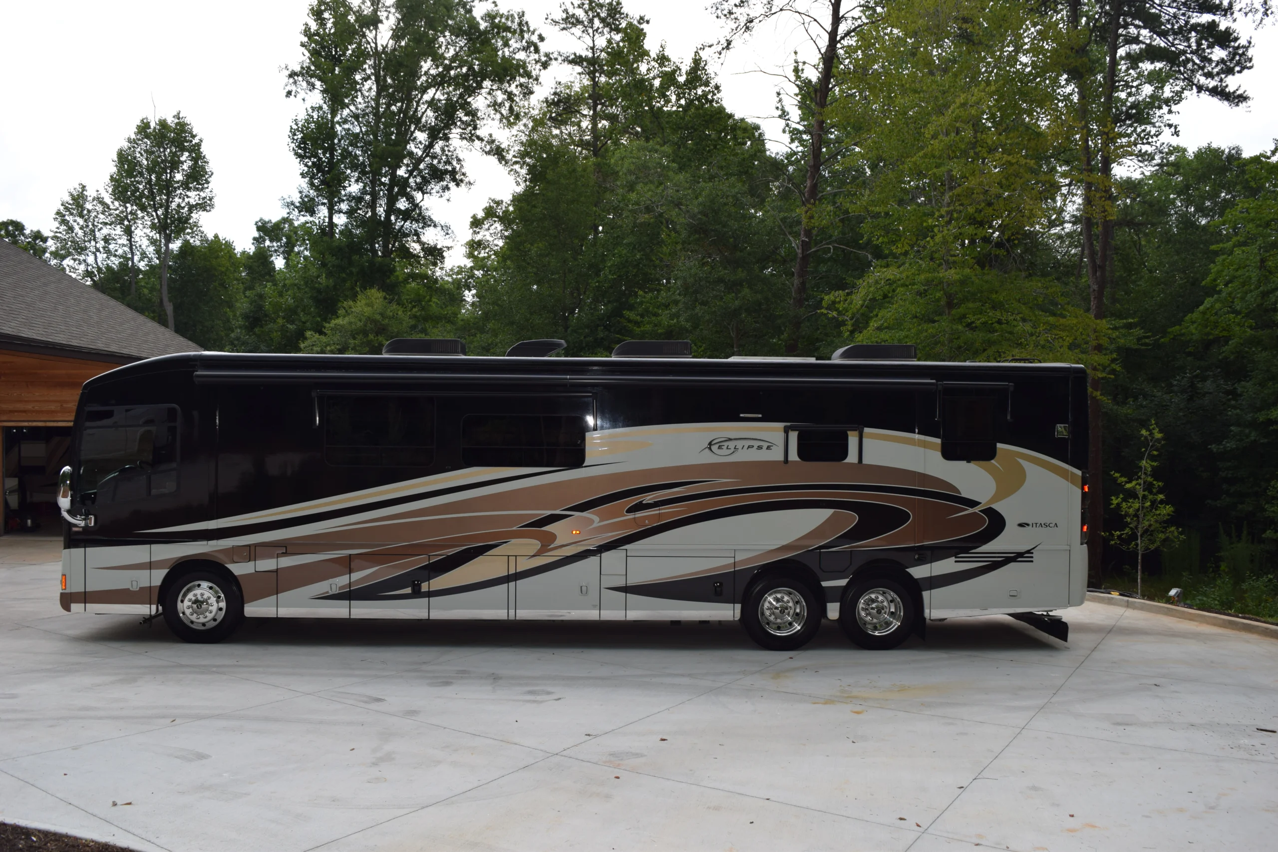 DSC_0530 A sleek motorcoach with elegant brown and beige swirl design parked on a driveway, surrounded by lush trees.