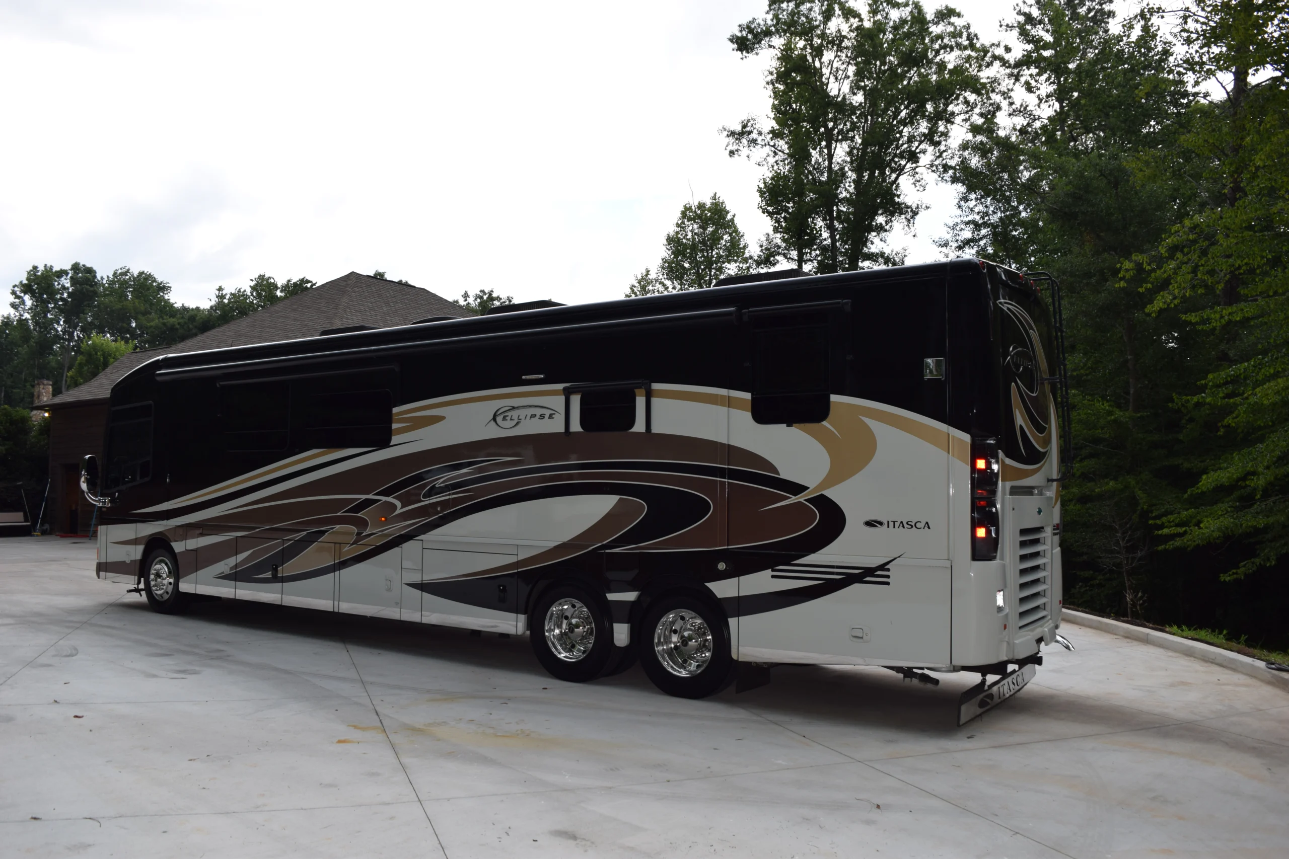 DSC_0531 Elegant Itasca motorcoach with brown and beige accents parked on a concrete driveway, surrounded by trees.