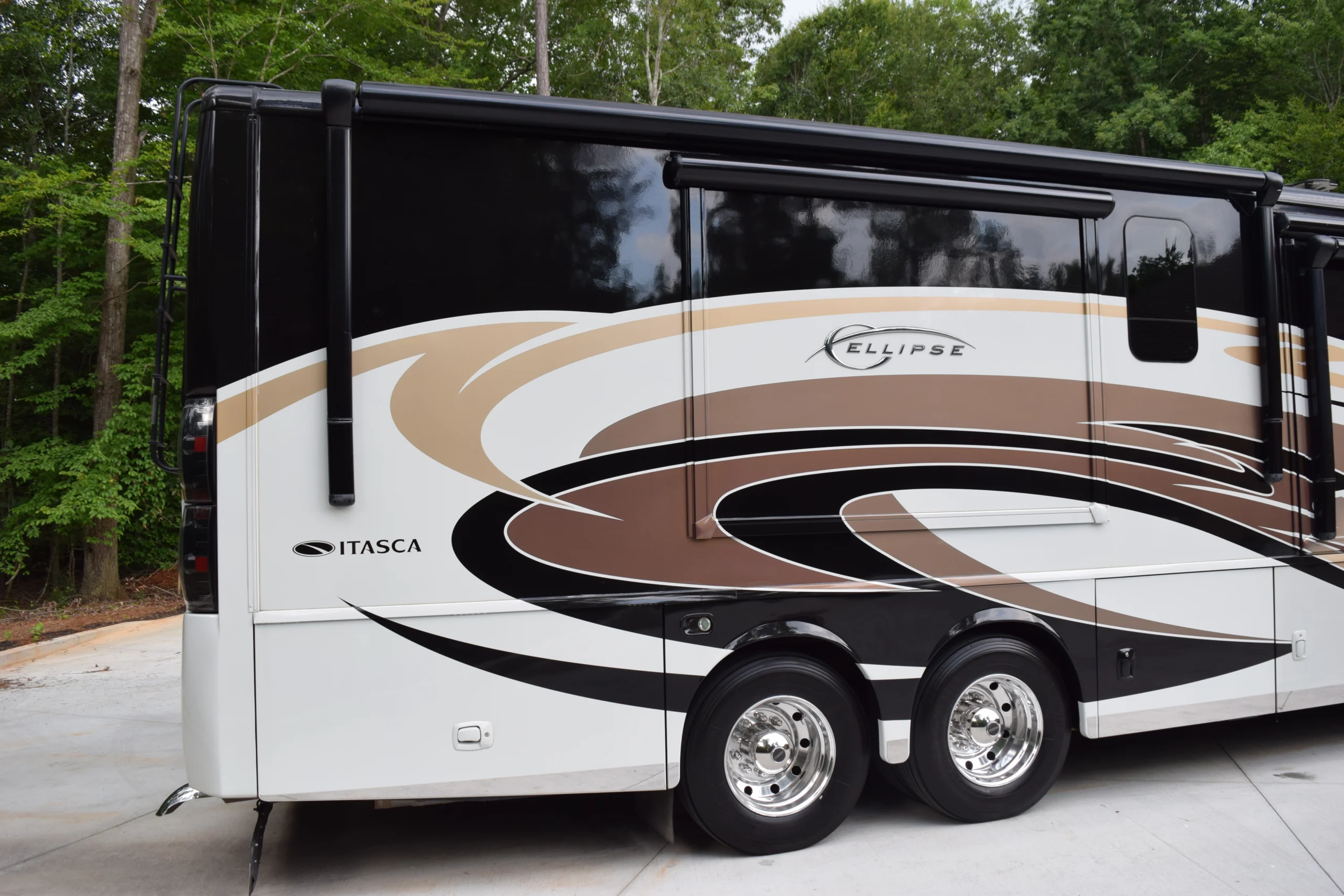 DSC_0532 Sleek black and white Itasca motorcoach with curved brown and beige designs, parked on a paved area near lush trees.