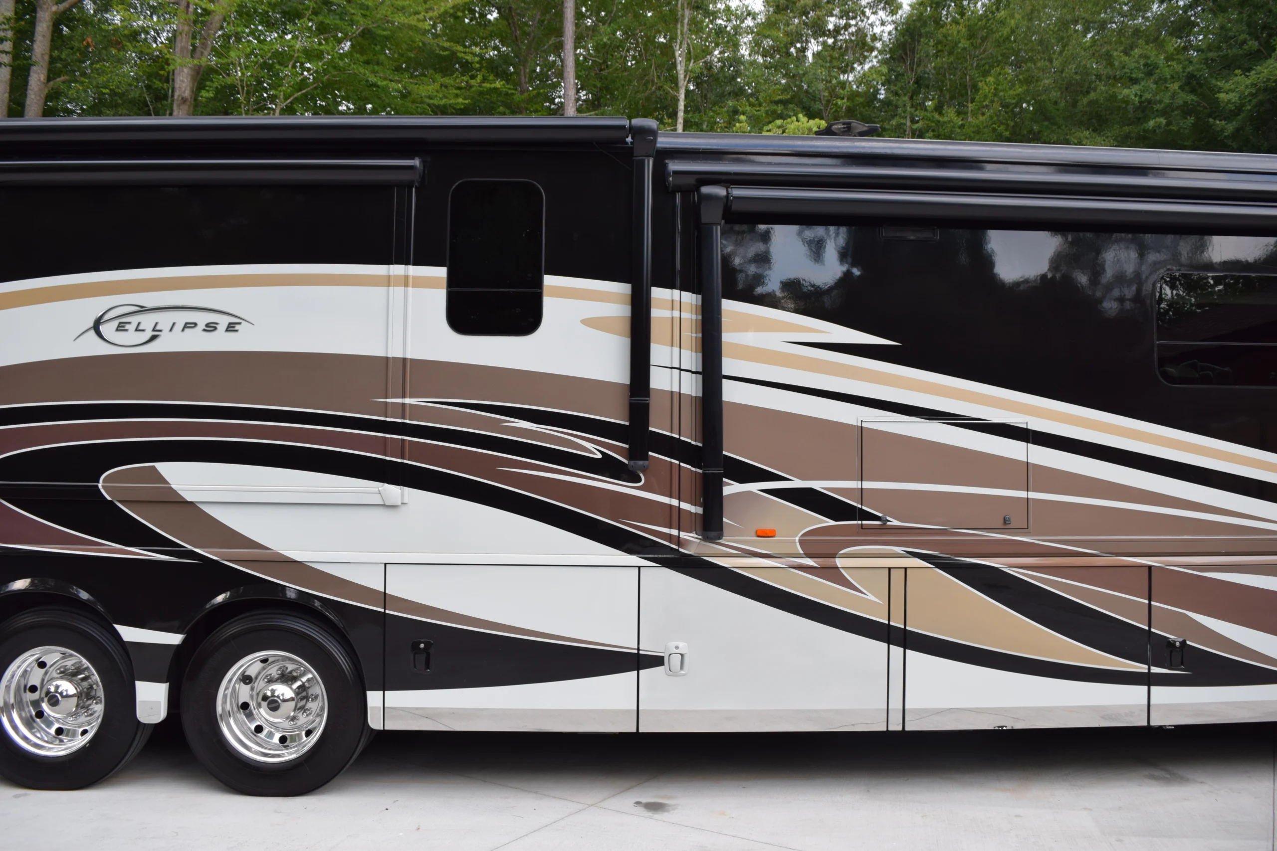 DSC_0533 Luxury motorcoach RV with sleek, curved brown and white designs, parked on concrete with trees in the background.