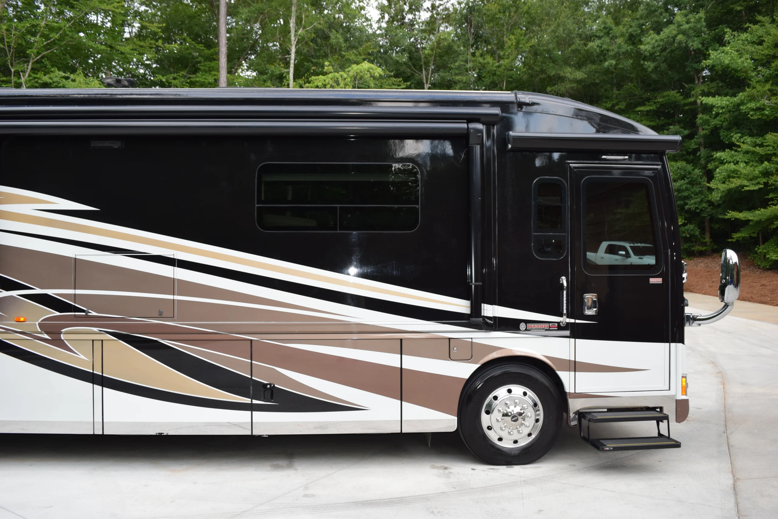 DSC_0534 Black motorcoach with brown, gold, and white stripes parked on concrete, surrounded by lush greenery.