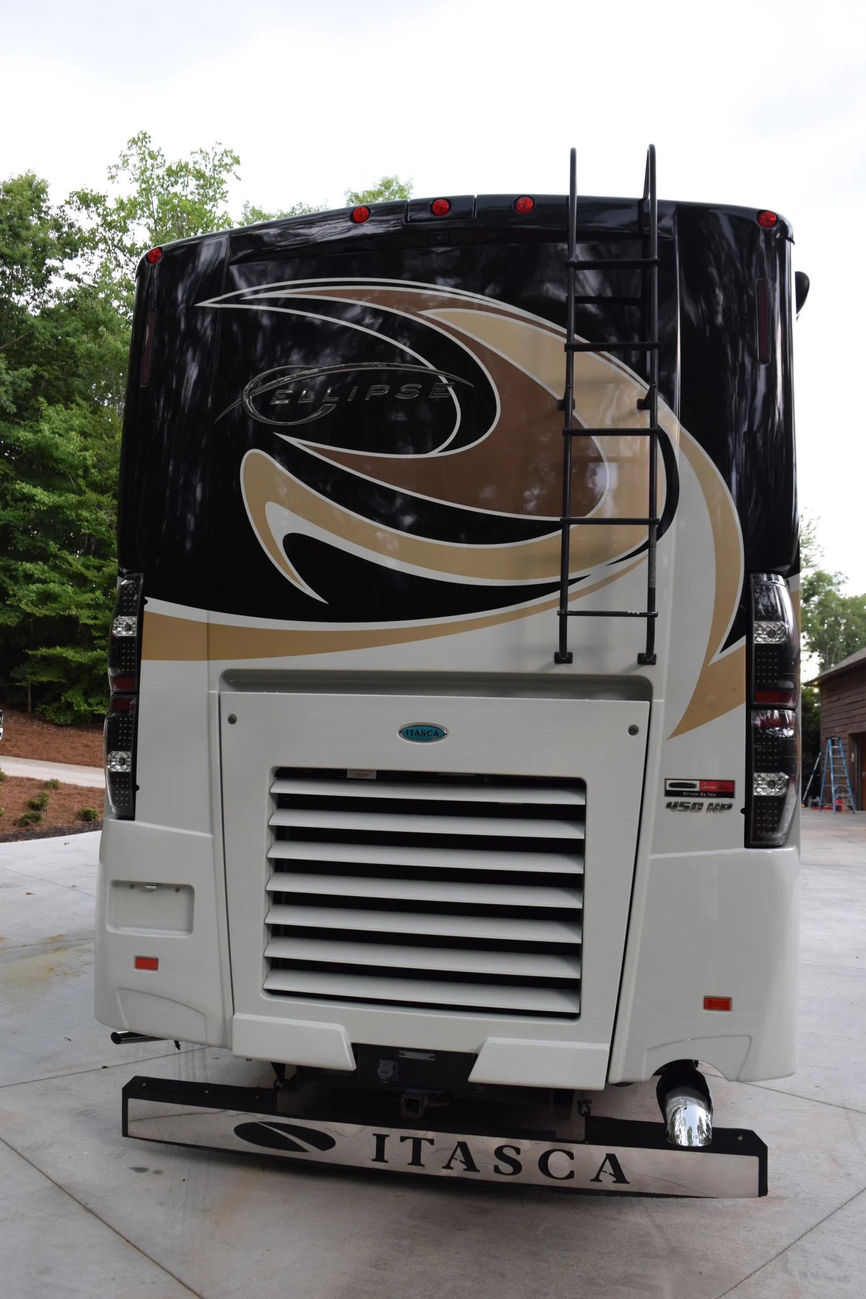 DSC_0535 Back of an Itasca motorcoach RV with a ladder, large vents, and stylish design, parked on a paved driveway surrounded by trees.