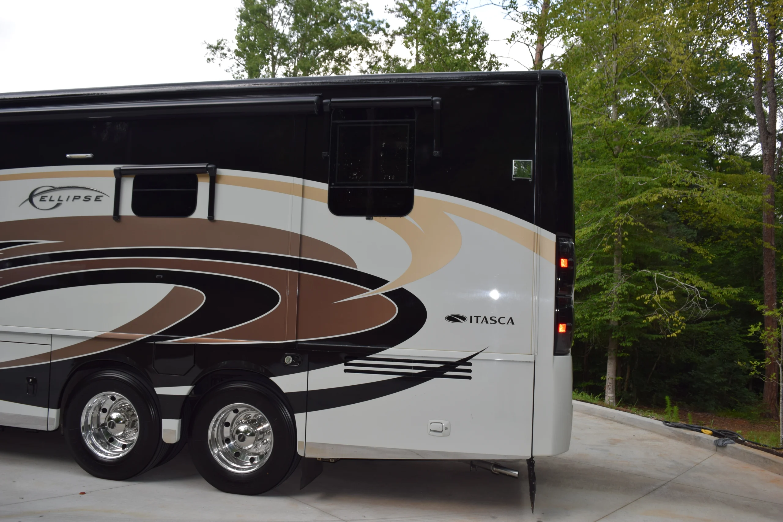 DSC_0538 Rear section of an Itasca motorcoach with decorative swirls, parked on a concrete driveway, surrounded by trees.