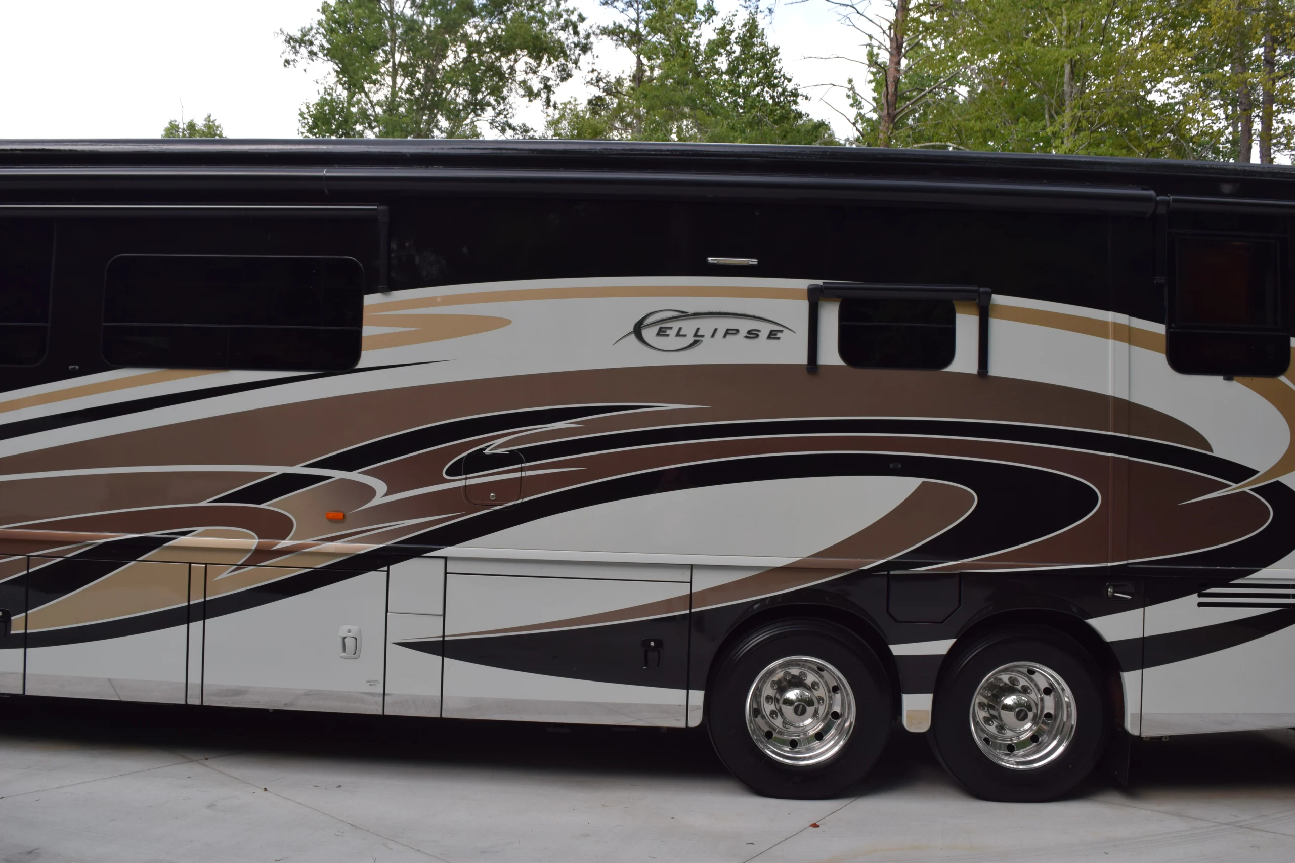 DSC_0540 Luxury motorcoach with sleek brown and black swirl patterns, parked on a concrete driveway amidst trees.
