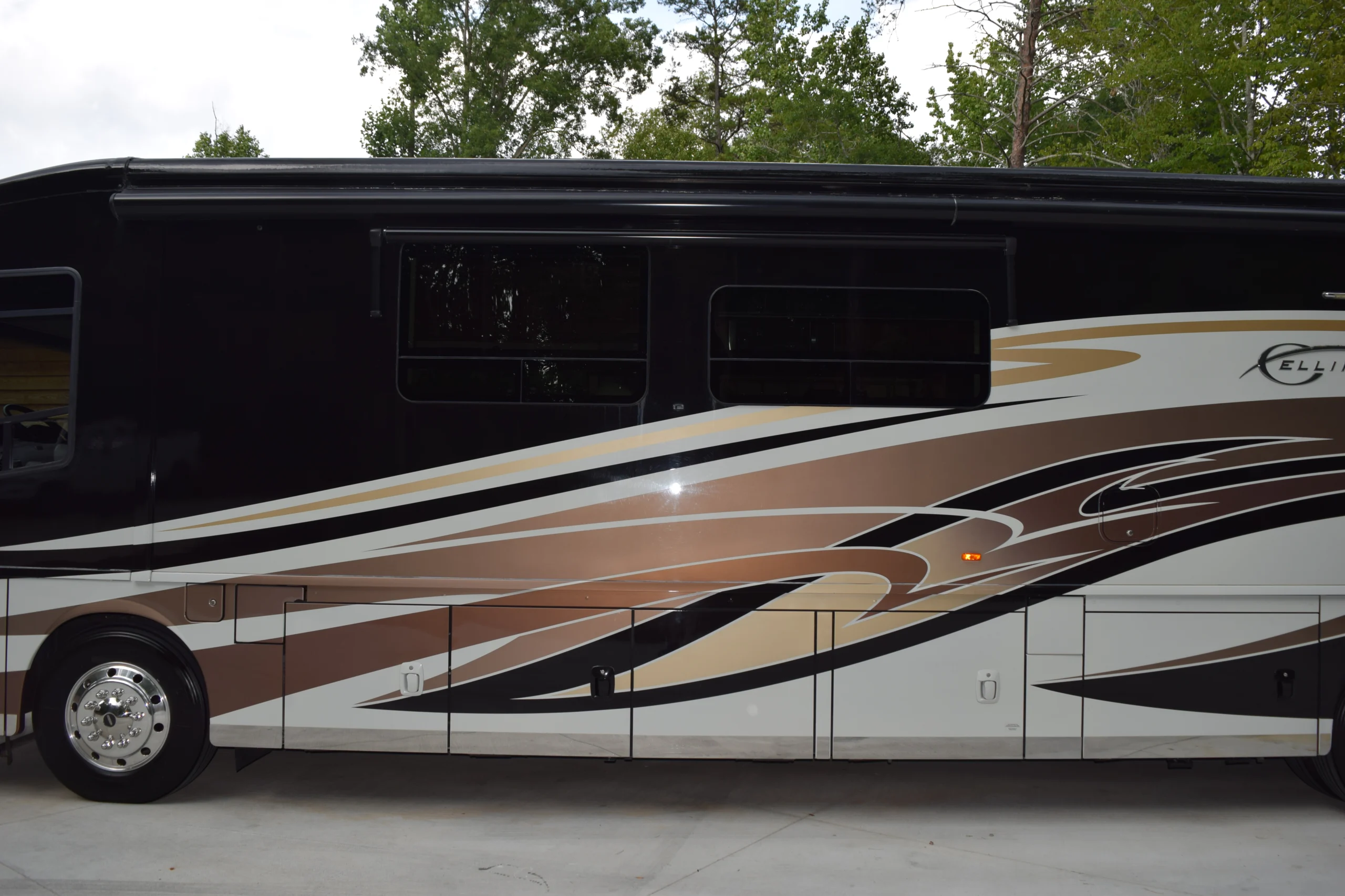 DSC_0541 Sleek motorcoach with brown, gold, and black swoosh design, parked on concrete beside a wooded area.
