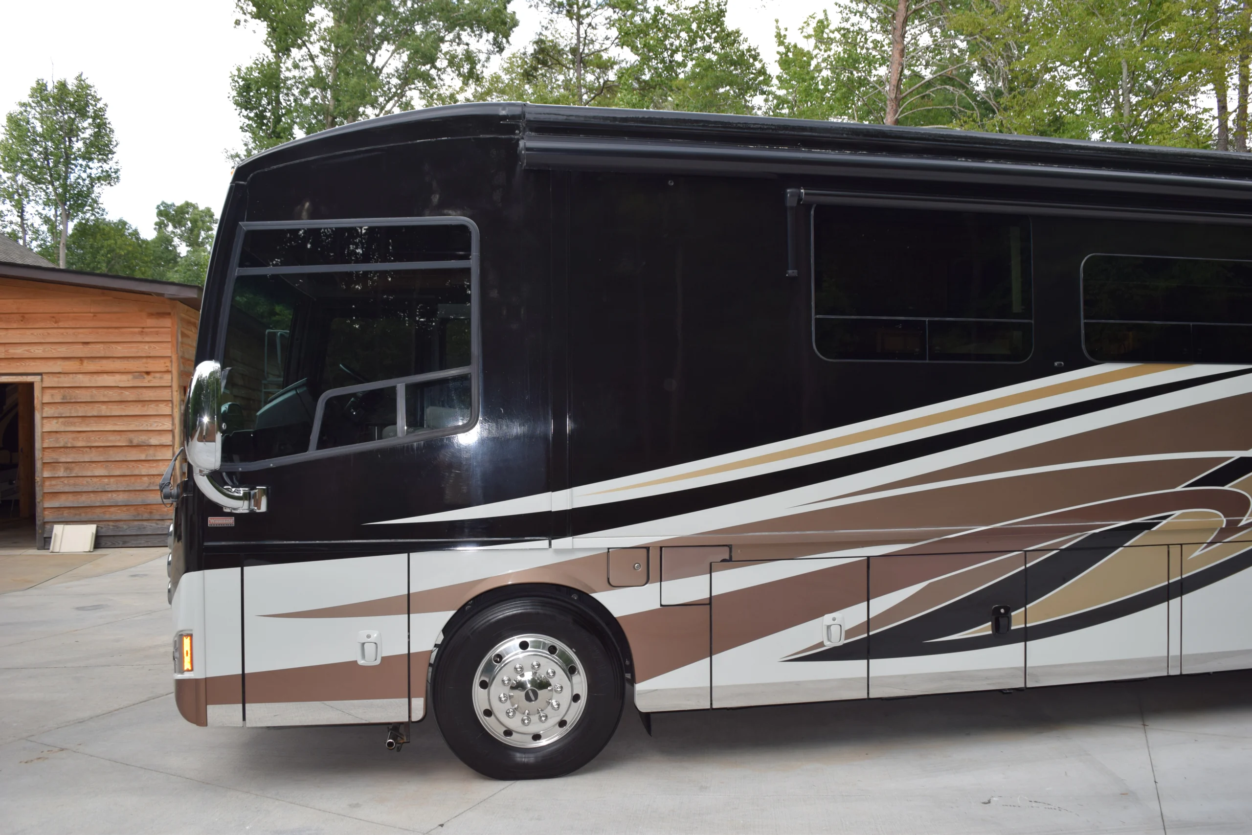 DSC_0542 Black, brown, and white motorcoach with sleek design parked near a wooden building in a wooded area.