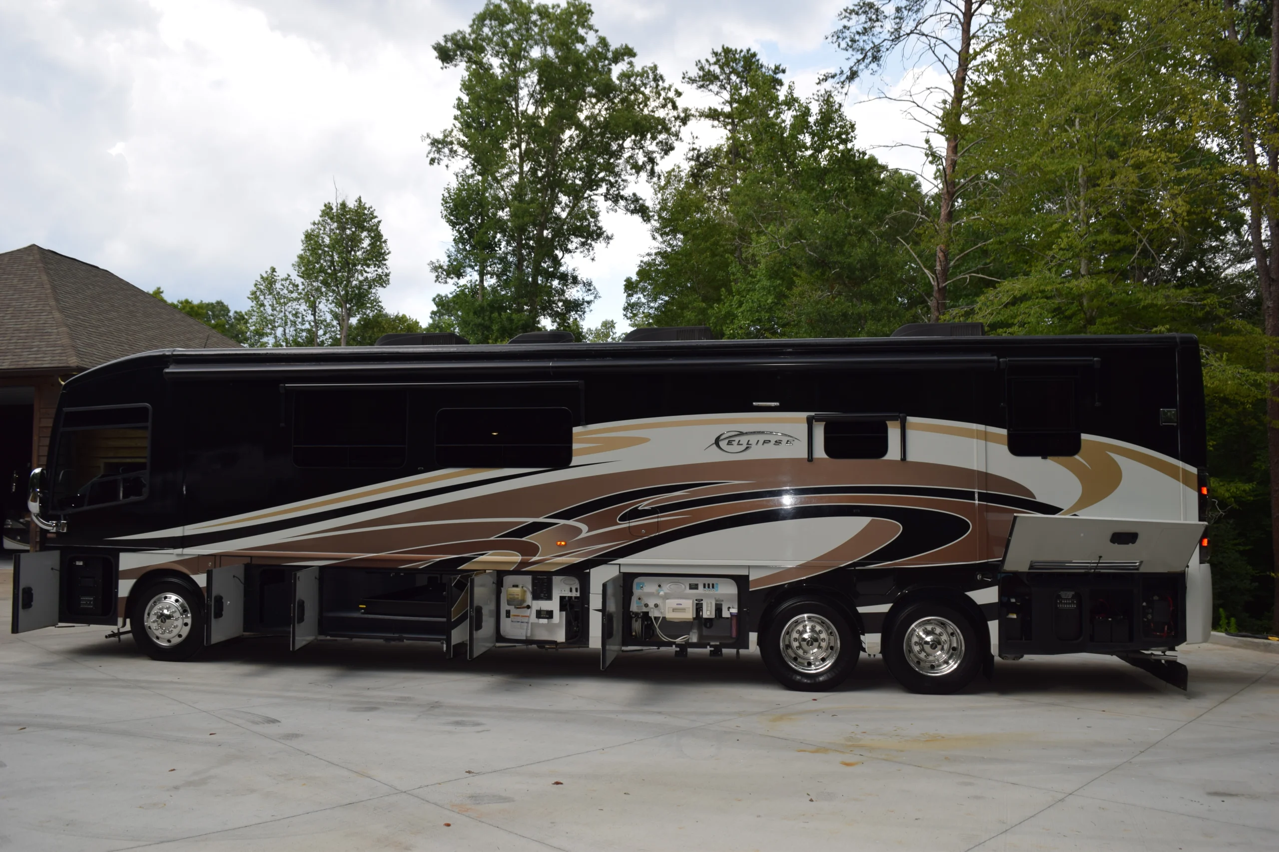 DSC_0543 Large Motorcoach with brown and black design, parked on a driveway with open storage compartments, surrounded by trees.