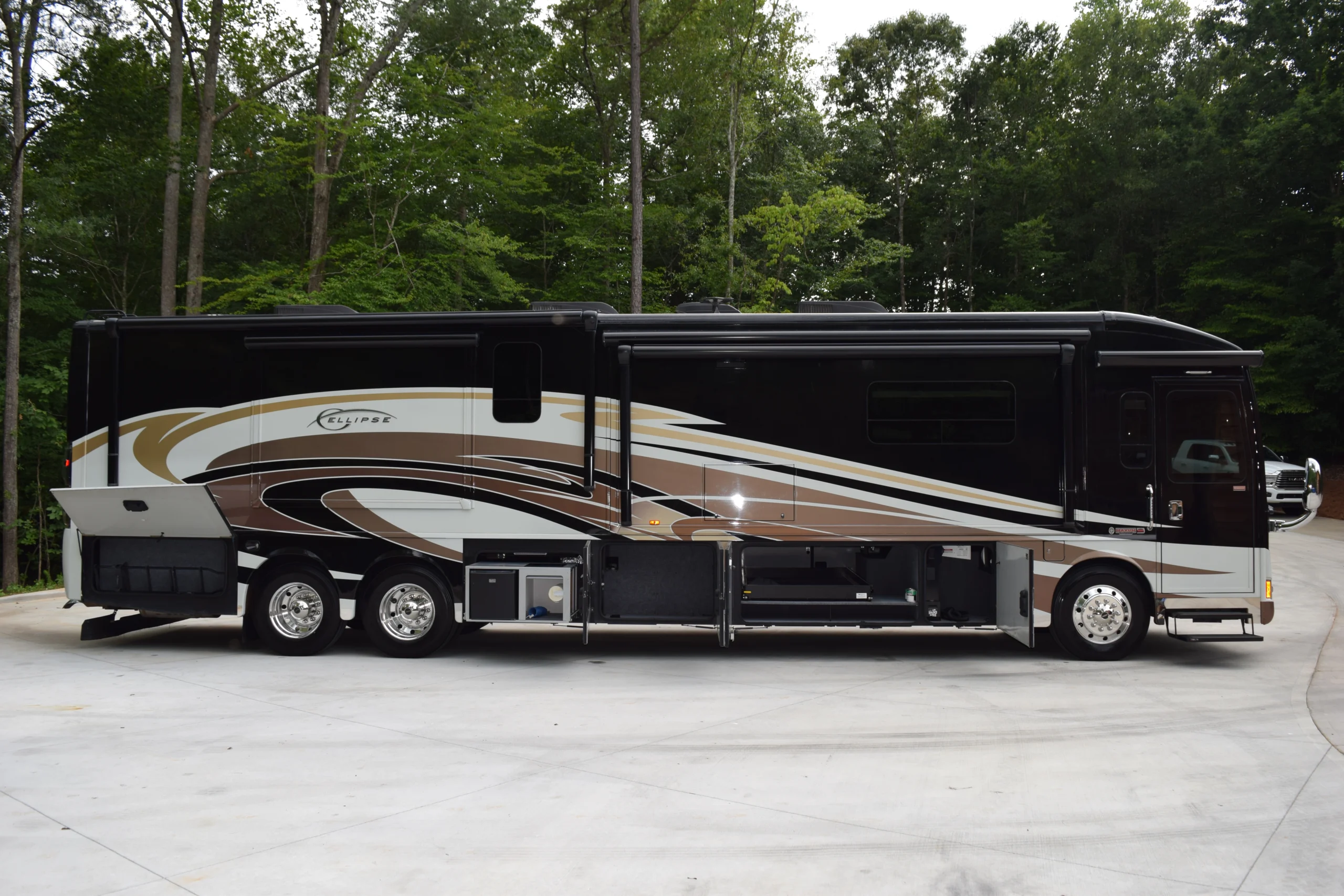 DSC_0545 Luxury motorcoach with sleek black, brown, and white design parked on concrete lot, surrounded by trees.