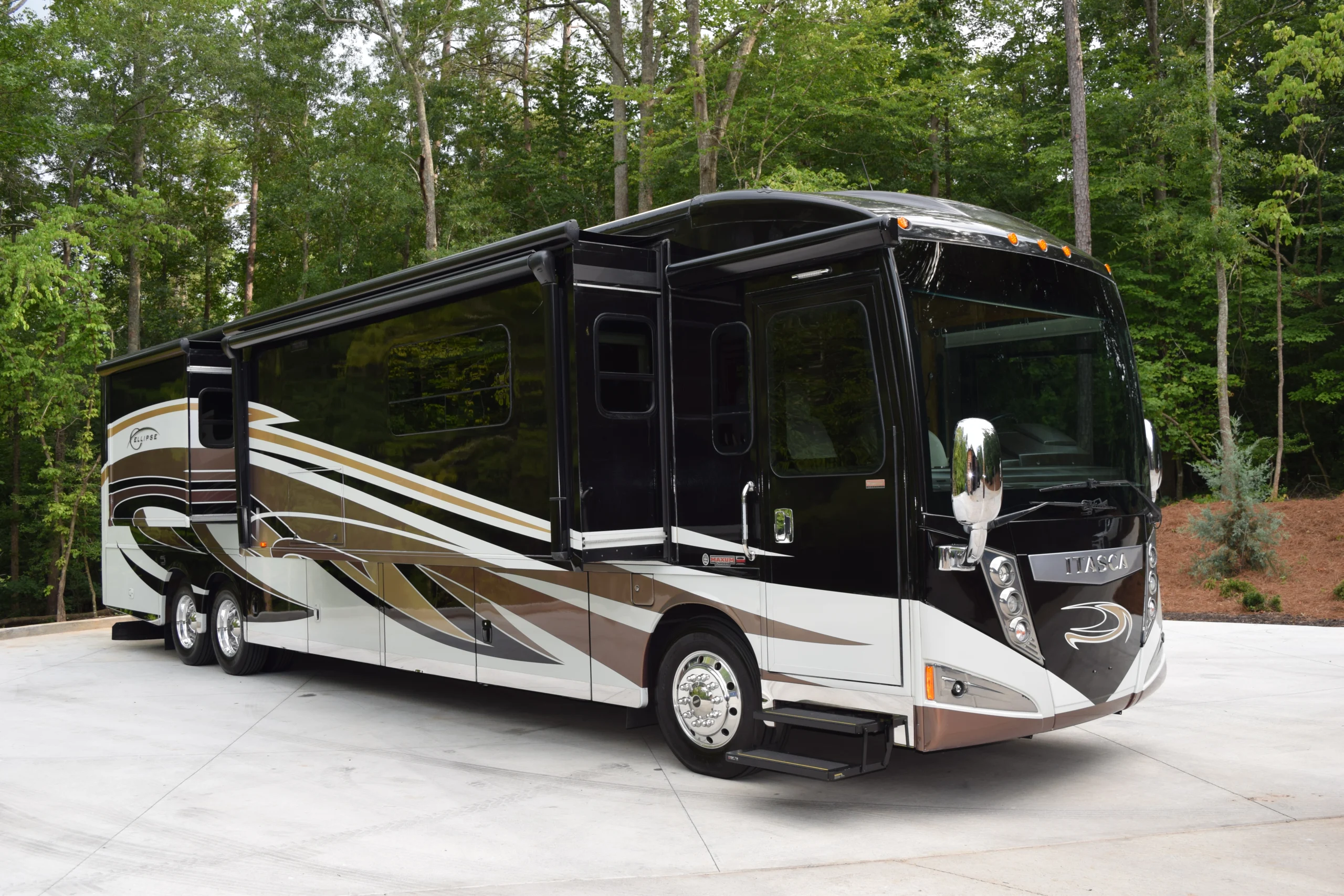 DSC_0546 Luxurious black and white motorcoach with slide-outs, parked on a concrete surface surrounded by lush green trees.