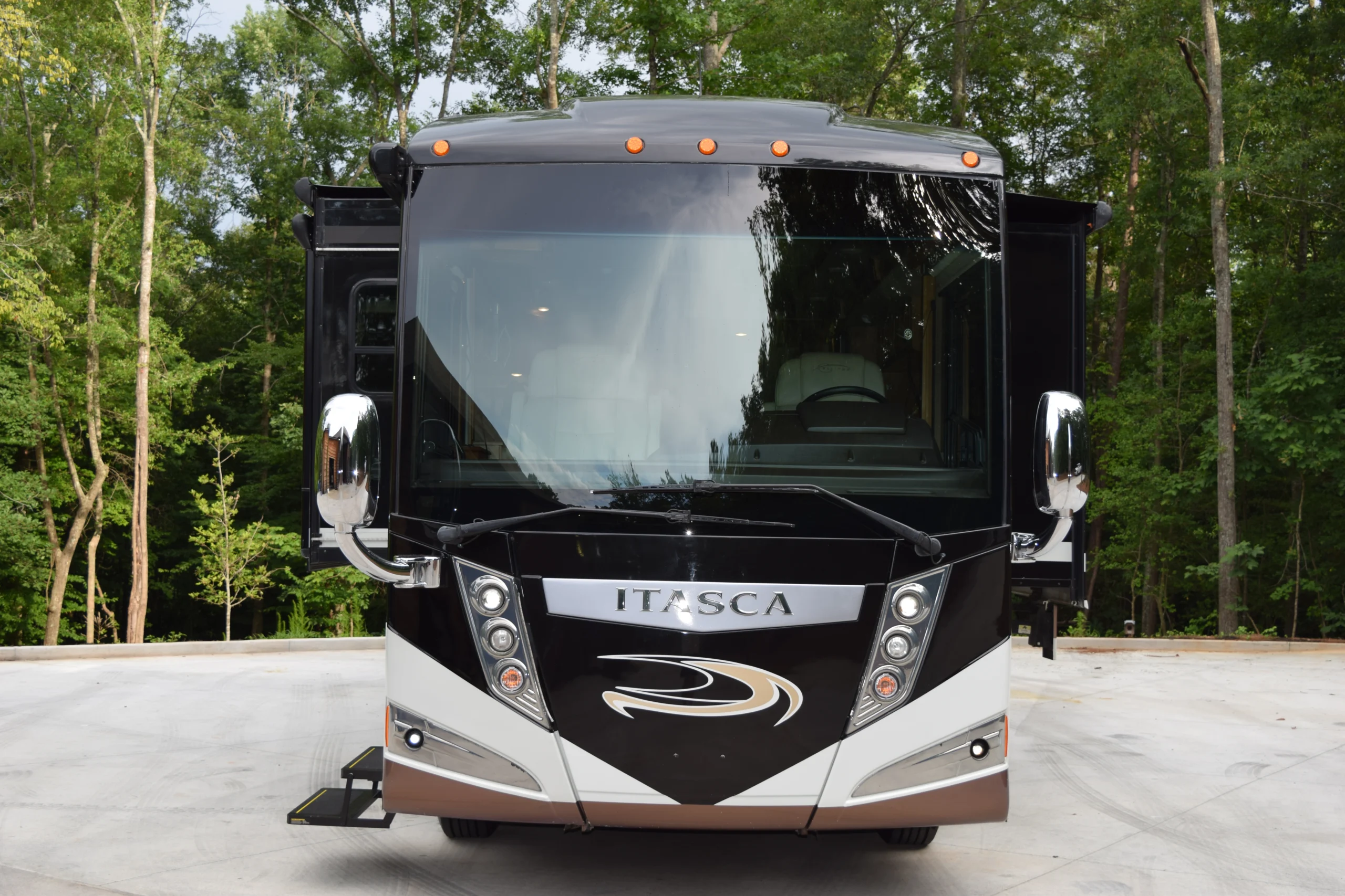 DSC_0547 Front view of a black Itasca motorcoach parked on a concrete surface, surrounded by lush green trees.