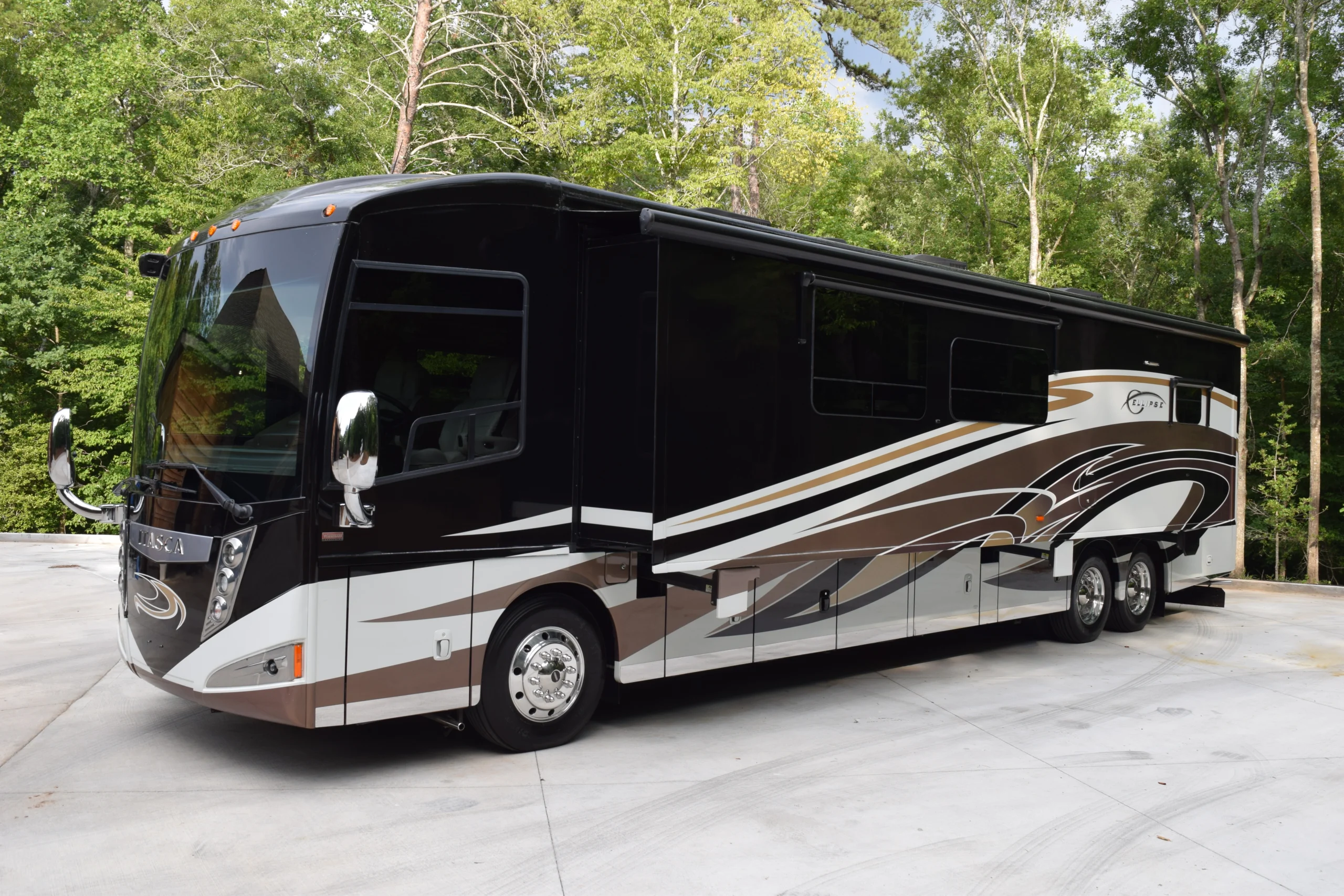 DSC_0550 Luxurious Motorcoach with sleek brown, white, and black design parked on concrete, surrounded by lush green trees.