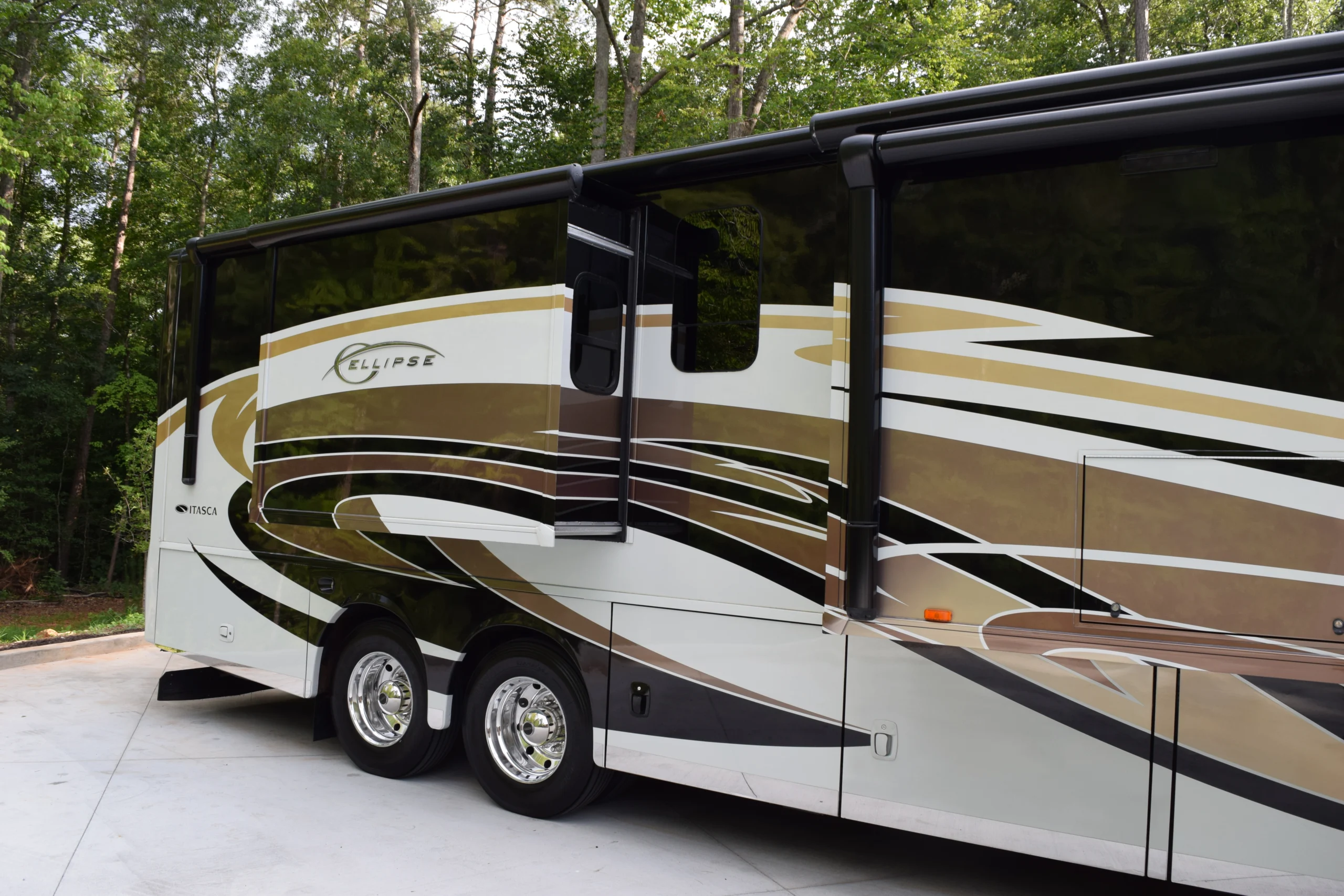 DSC_0554 Luxury motorcoach with slide-out extended, parked on concrete, surrounded by lush green trees in a forest setting.