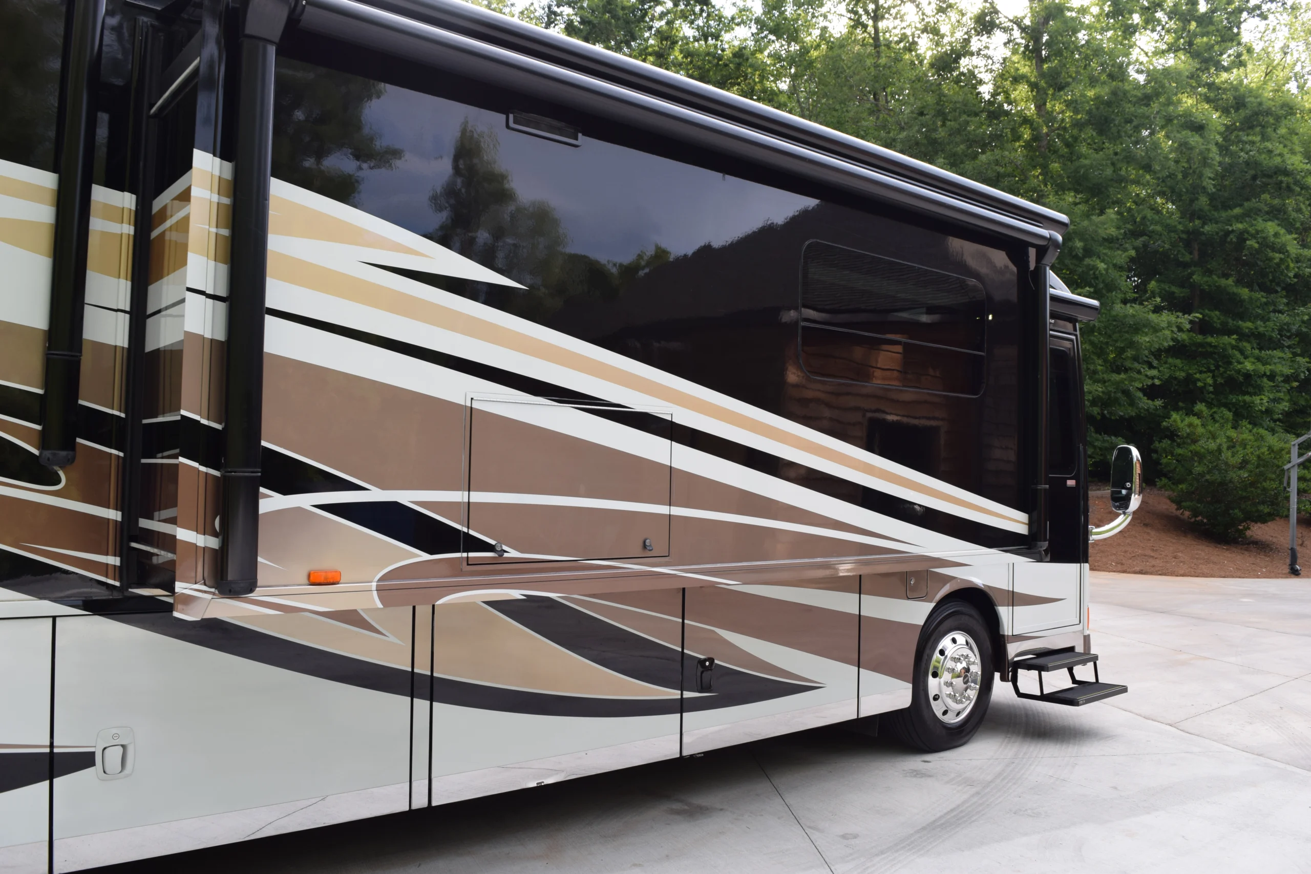 DSC_0555 Sleek motorcoach with brown and beige geometric design parked on a paved area, surrounded by lush greenery.