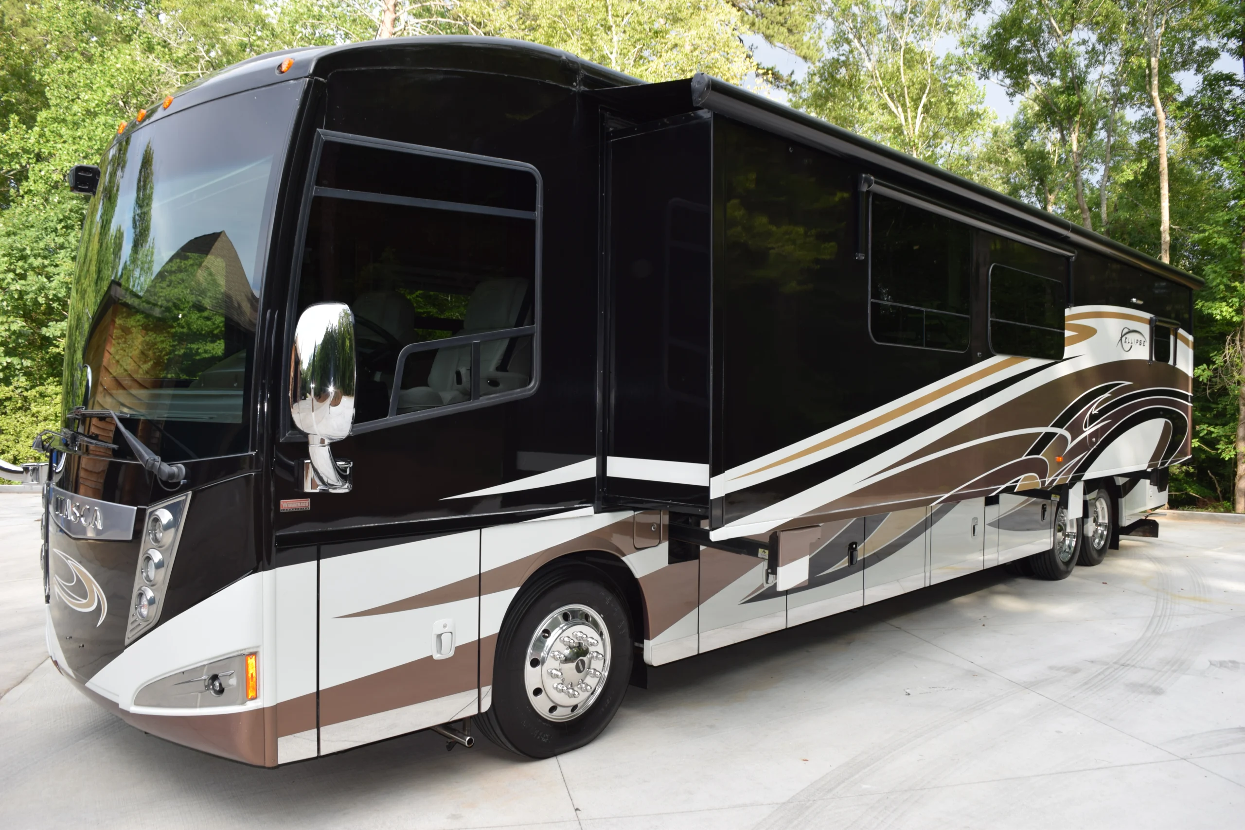 DSC_0556 Sleek black and white motorcoach with brown accents parked on a concrete driveway, surrounded by lush green trees.