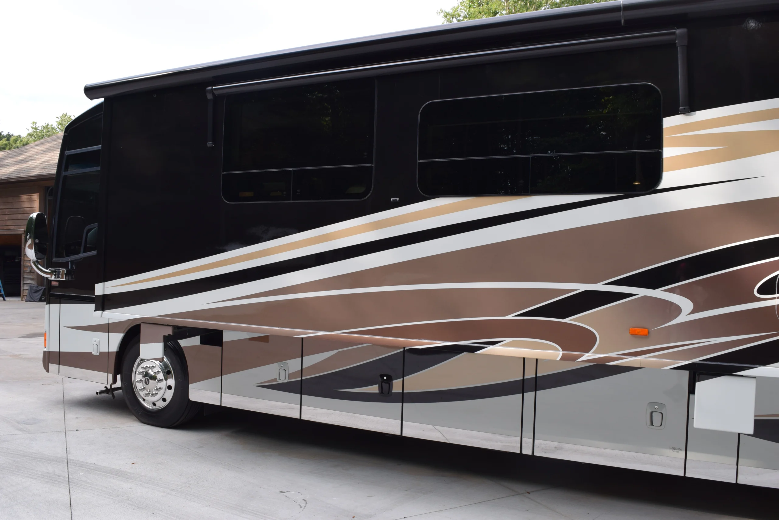 DSC_0557 Sleek Motorcoach with brown, black, and white stripes parked on a paved driveway, showcasing shiny silver wheels.