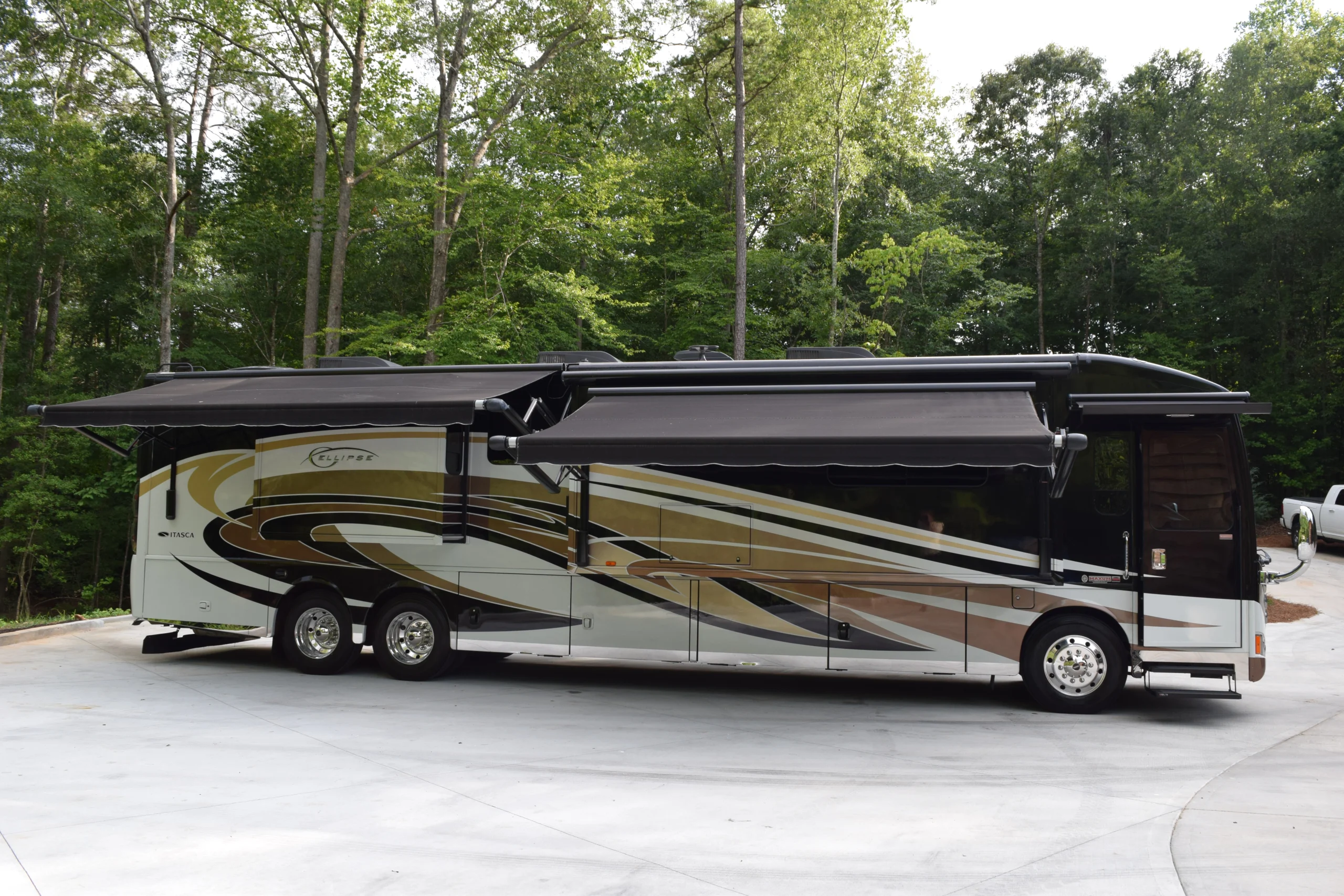 DSC_0560 Luxury motorcoach with extended awnings parked on a driveway, surrounded by lush green trees in the background.