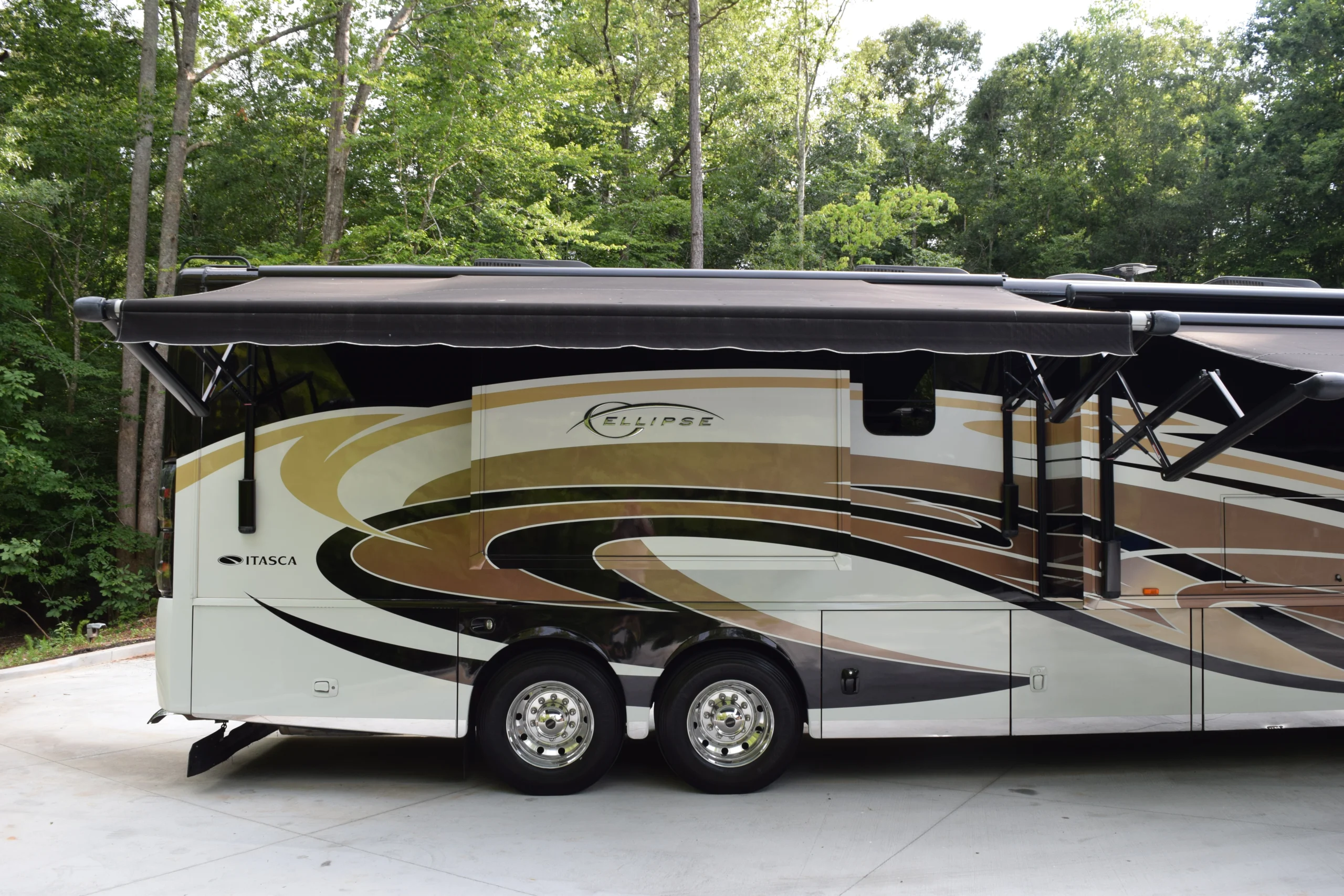 DSC_0561 Luxury motorcoach with retractable awning parked in a wooded area. Stylish brown and gold design on the side.