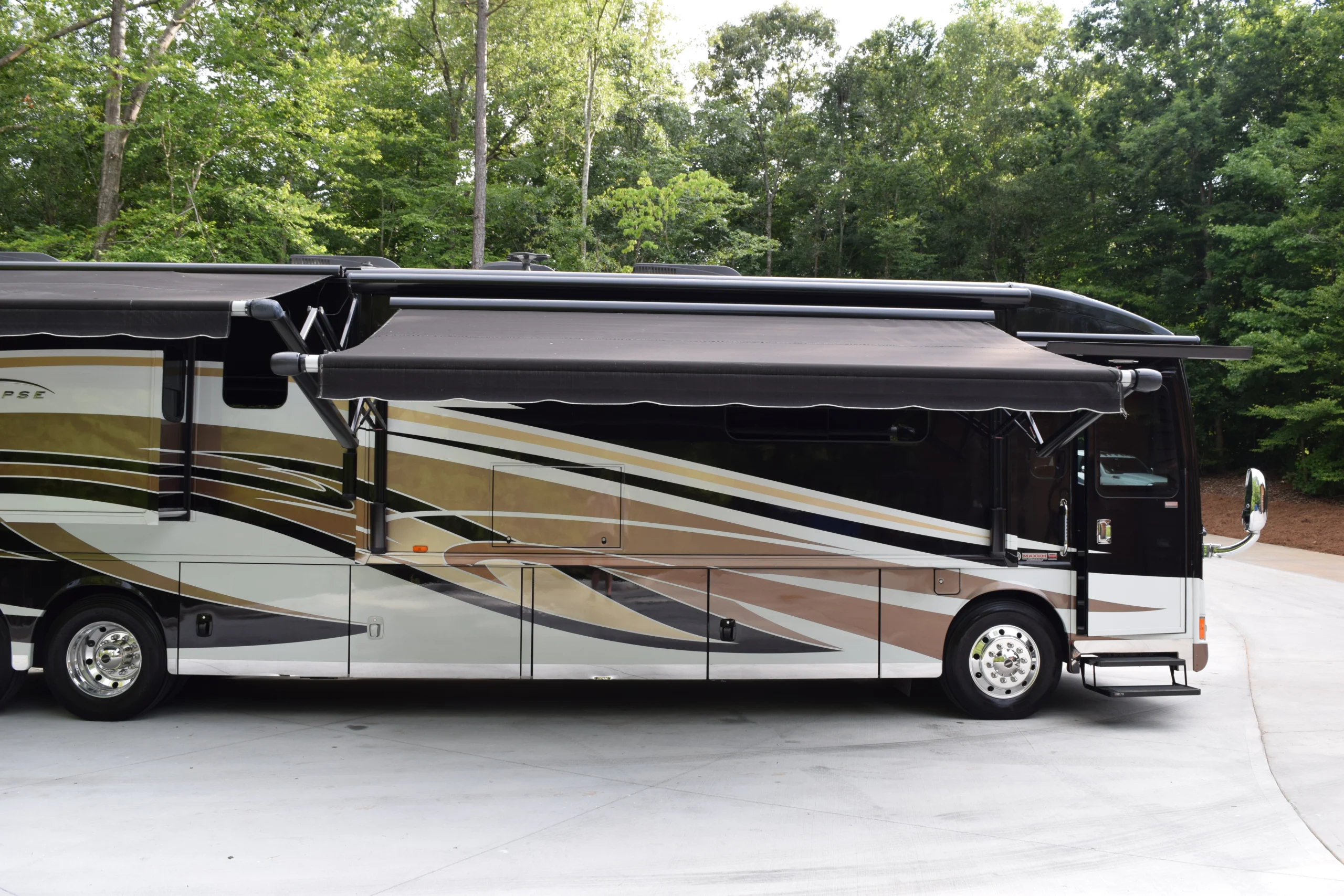 DSC_0562 Motorcoach with extended awning parked on a driveway, surrounded by lush green trees.