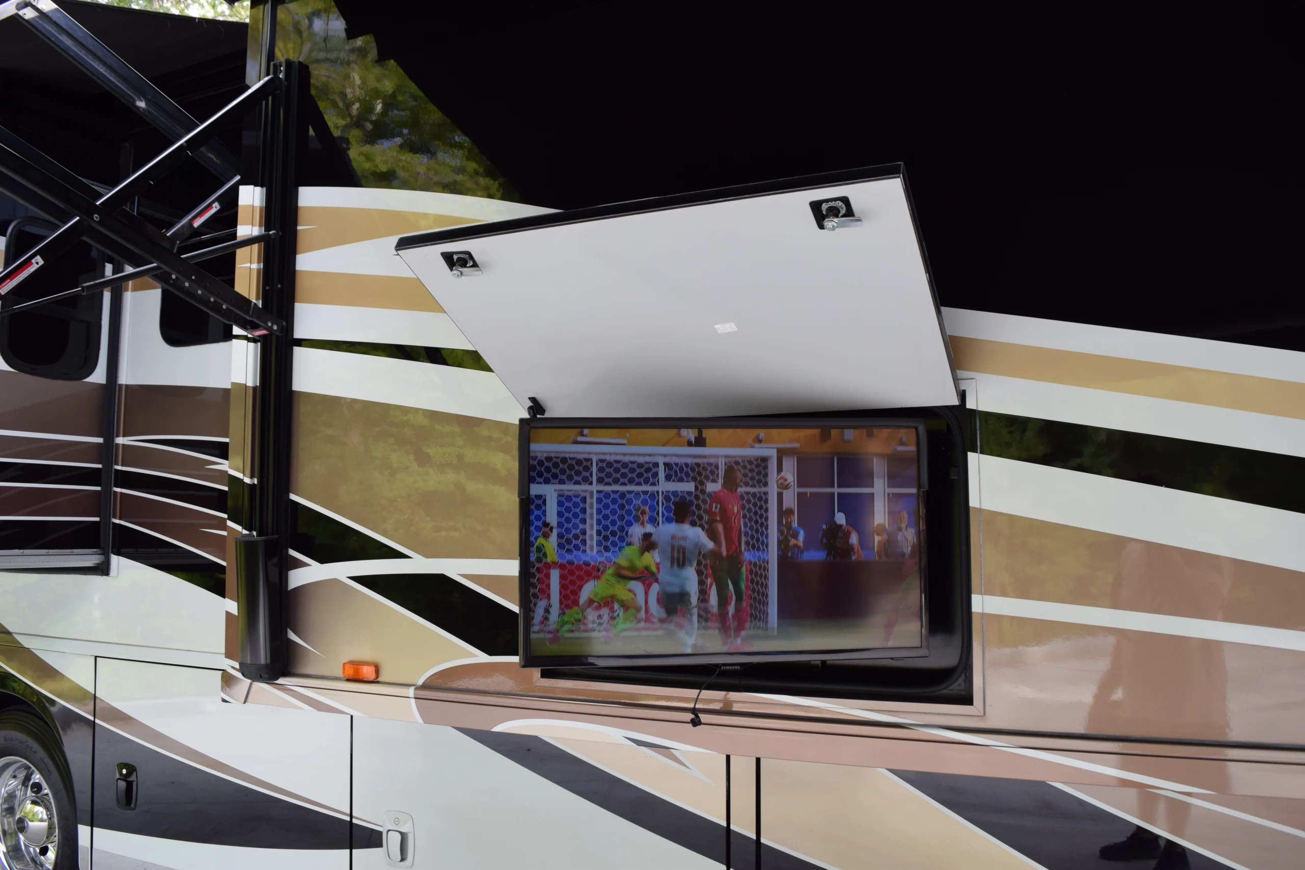 DSC_0564 Outdoor TV compartment on a motorcoach and RV showing a soccer match, surrounded by beige and brown exterior design.