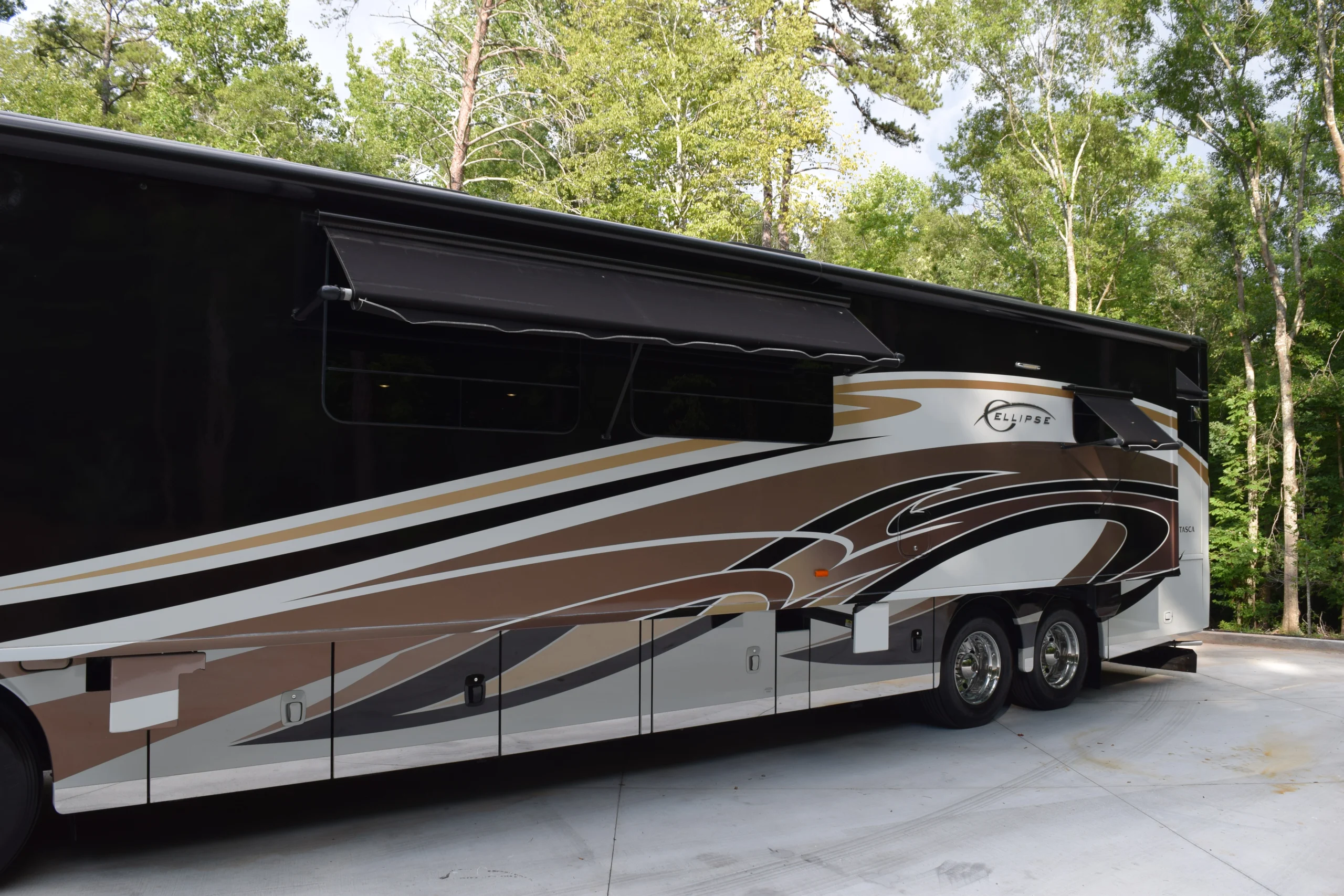 DSC_0565 Luxurious motorcoach with sleek, modern design featuring brown and beige swirls, parked on a driveway with a scenic forest backdrop.