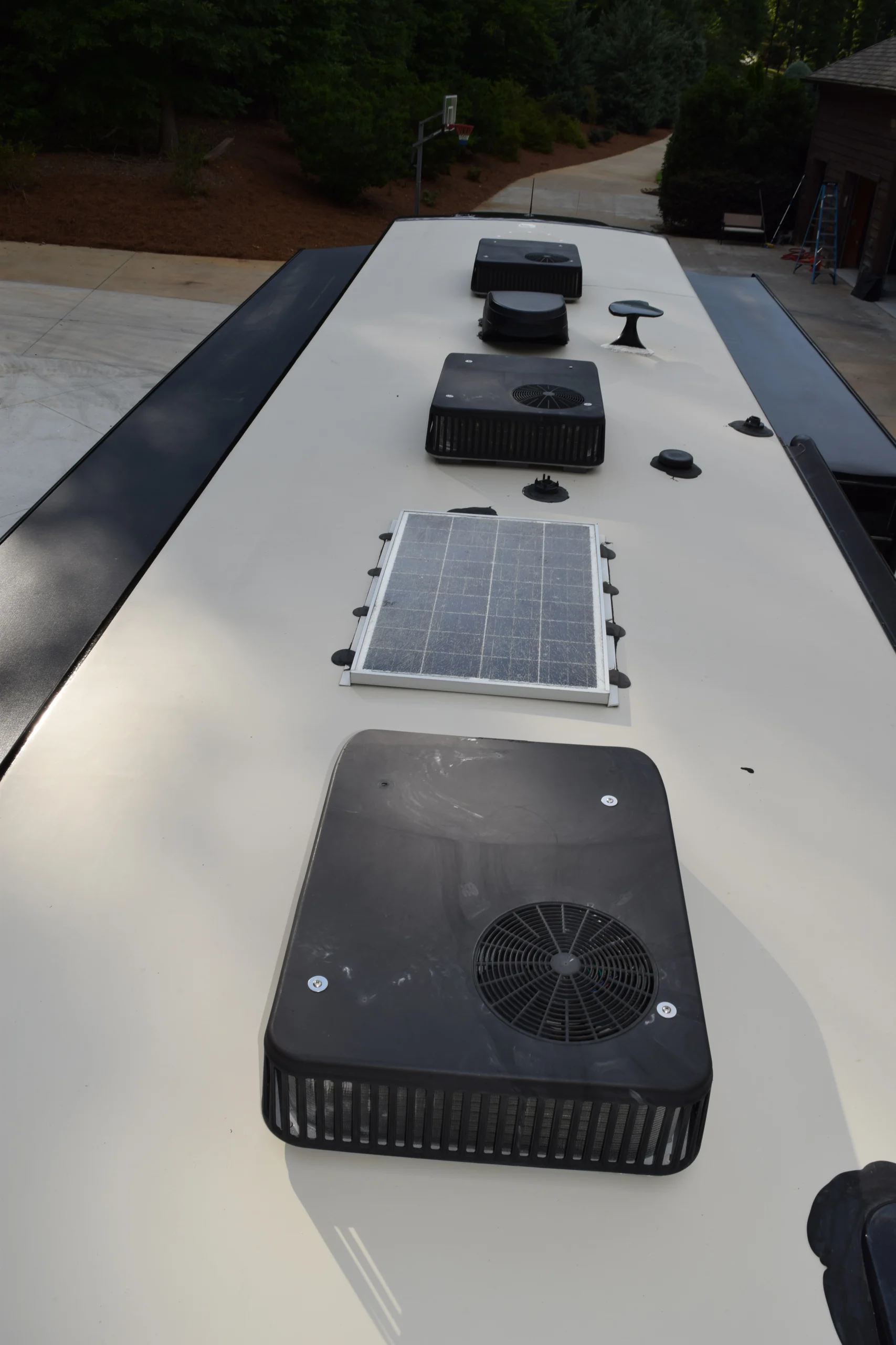 DSC_0570 Rooftop view of a Motorcoach and Rvs with solar panel, vents, and fans, surrounded by trees and a basketball hoop in the background.
