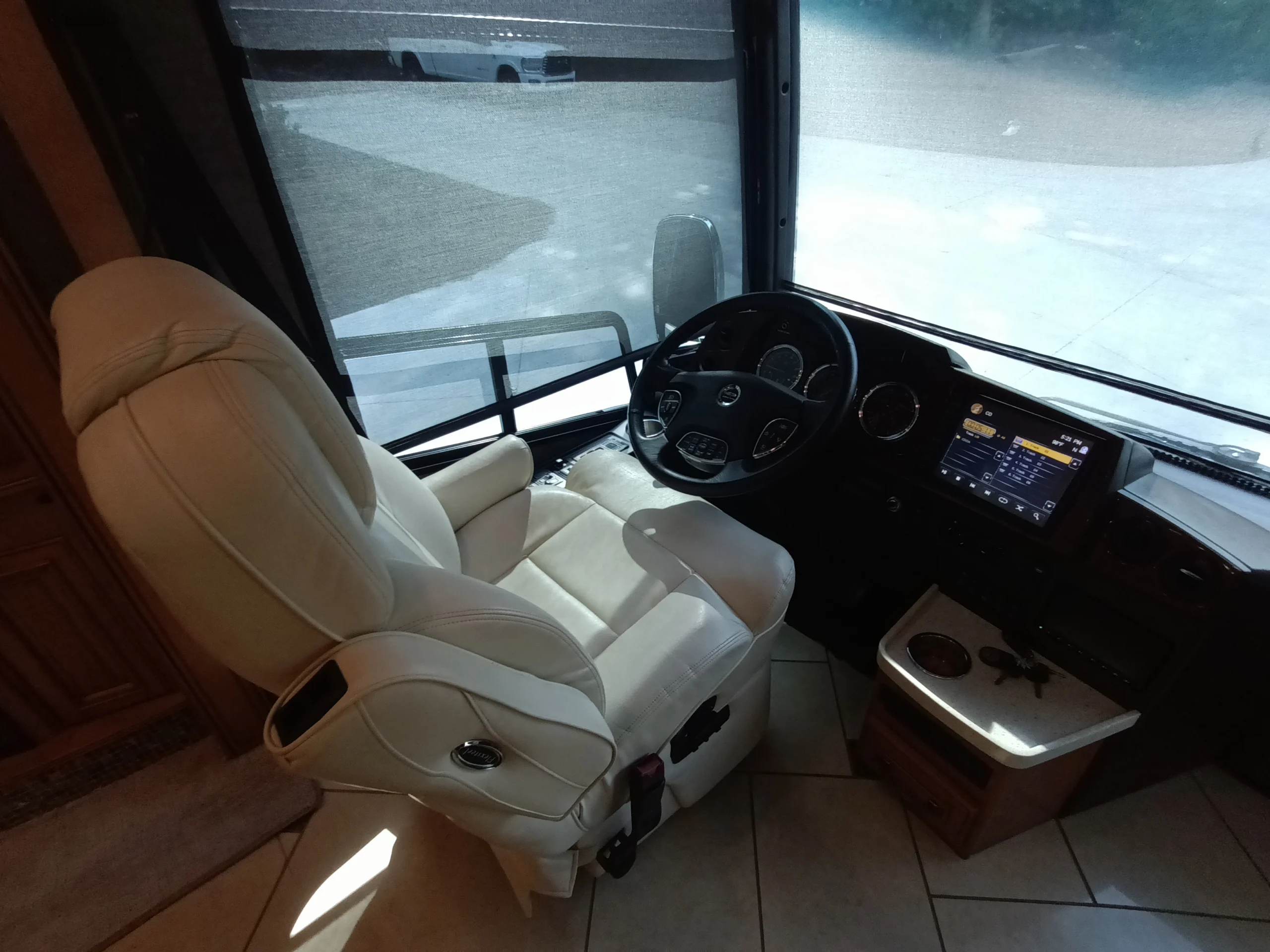 IMG_20260710_172039_626 Driver's seat in a Motorcoach and Rvs with a steering wheel, dashboard screen, and beige leather chair, overlooking a front window.