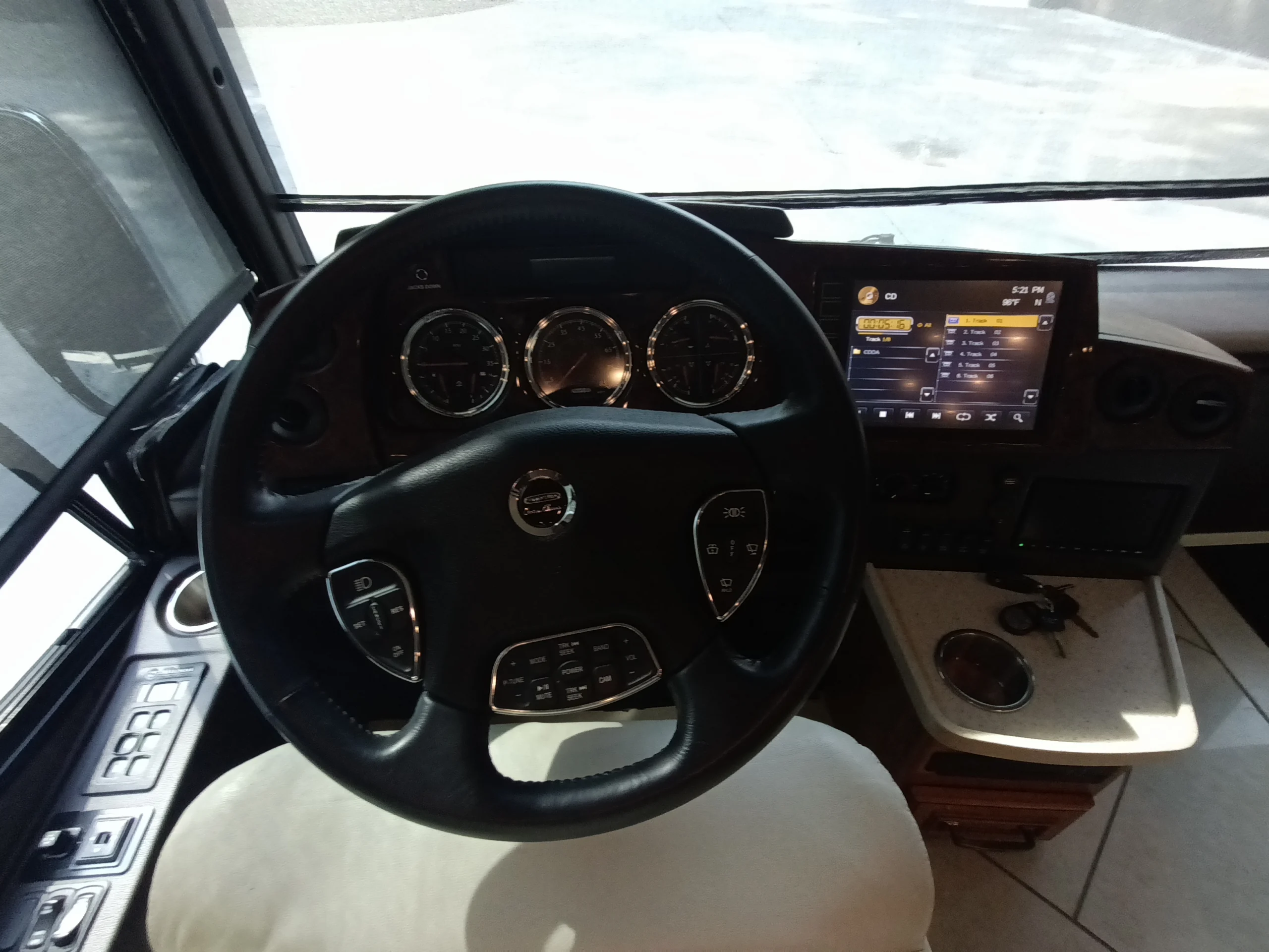 IMG_20260710_172044_649 Motorcoach dashboard with steering wheel, gauges, and touchscreen displaying radio. Armrest with cup holder and keys on the side.