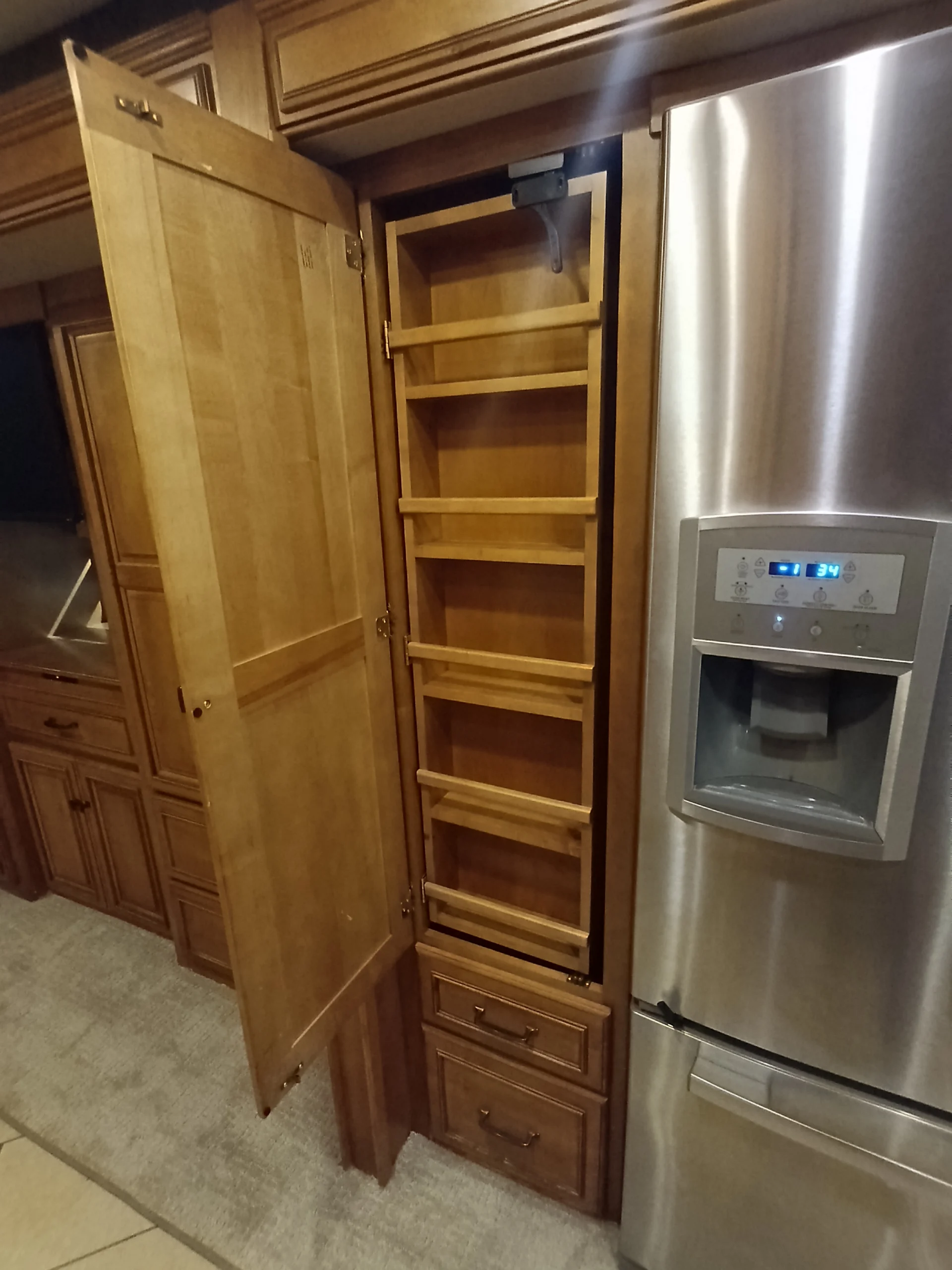 IMG_20260710_172339_017 Wooden pull-out pantry beside a stainless steel fridge, within a Motorcoach and Rvs interior.
