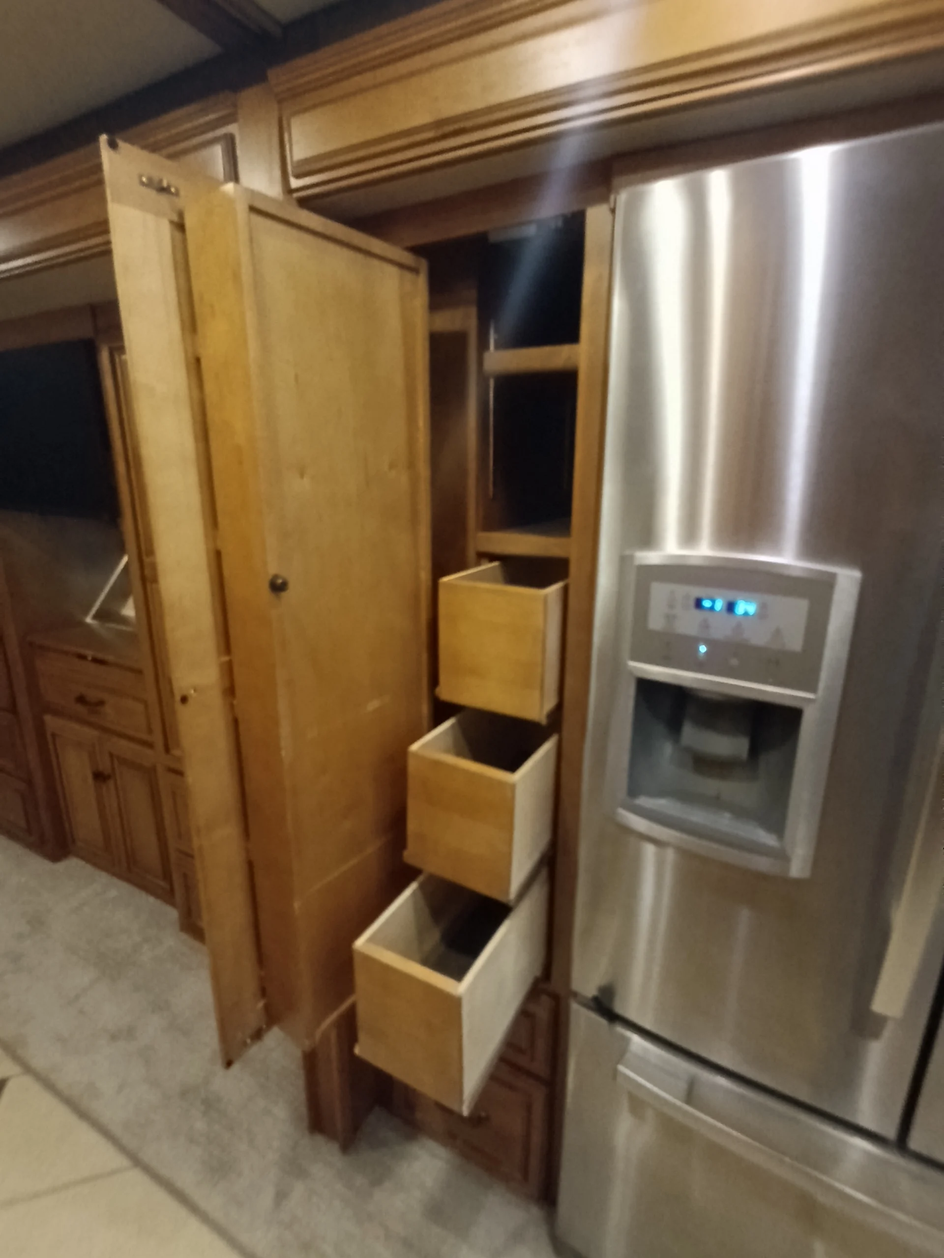 IMG_20260710_172351_482 Wooden pull-out pantry drawers next to a stainless steel refrigerator in a motorcoach and RVs kitchen area.