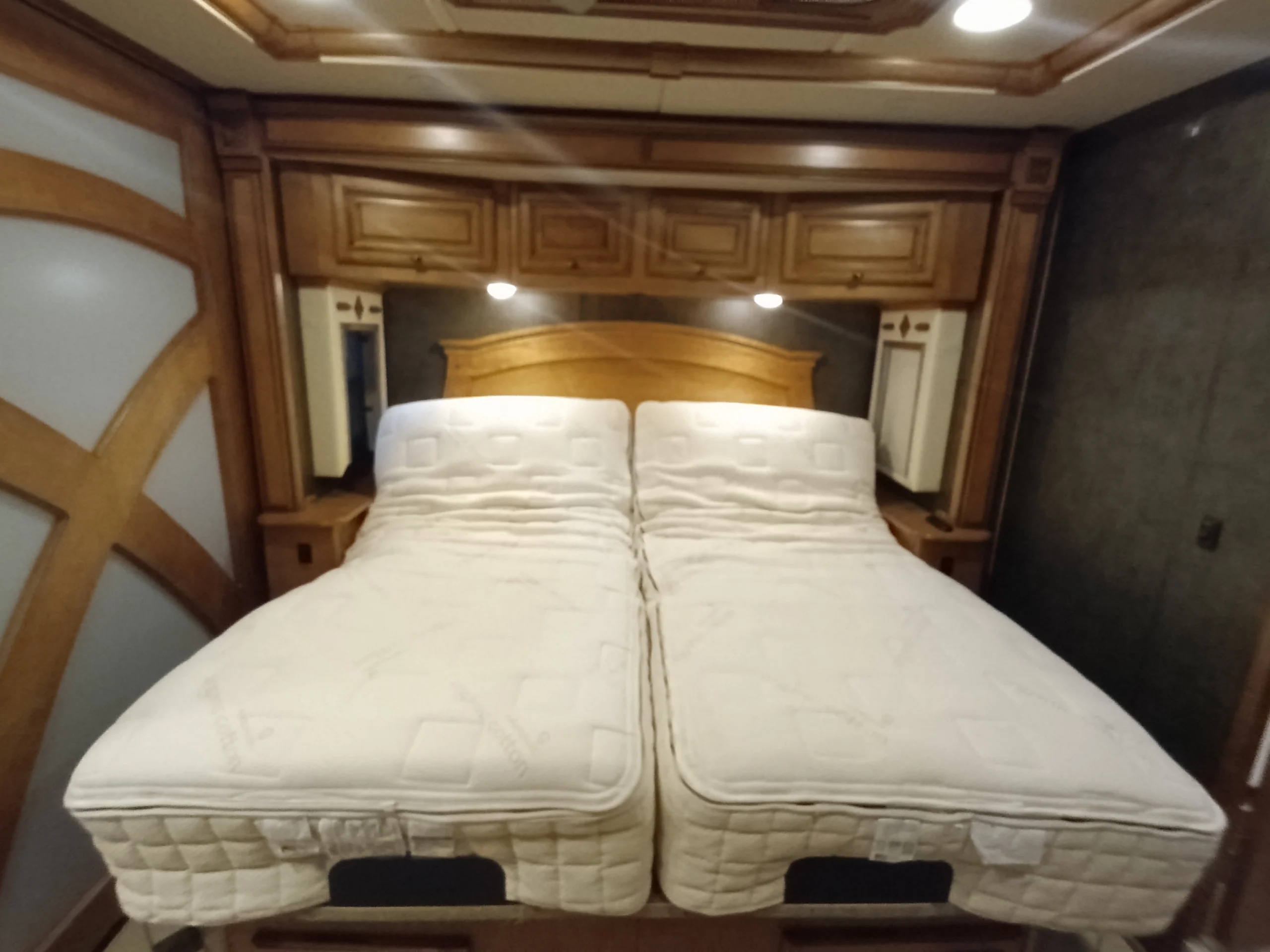 IMG_20260710_172559_802 Twin adjustable beds with quilted mattresses in a motorcoach and RVs interior, featuring wooden cabinetry and soft lighting.