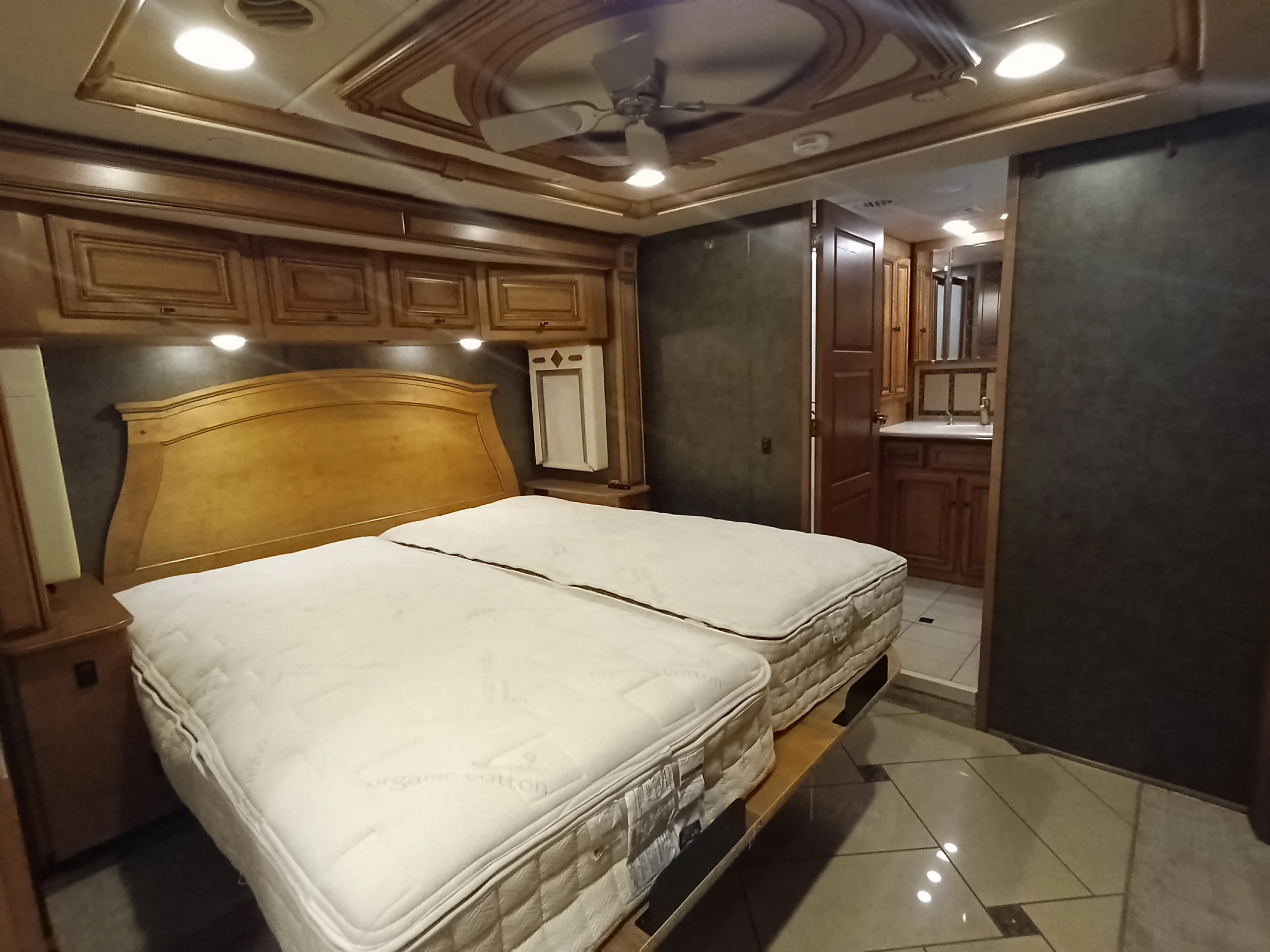 IMG_20260710_172648_716 Spacious motorcoach interior with twin beds, overhead storage, elegant wood paneling, and a view into a private bathroom.