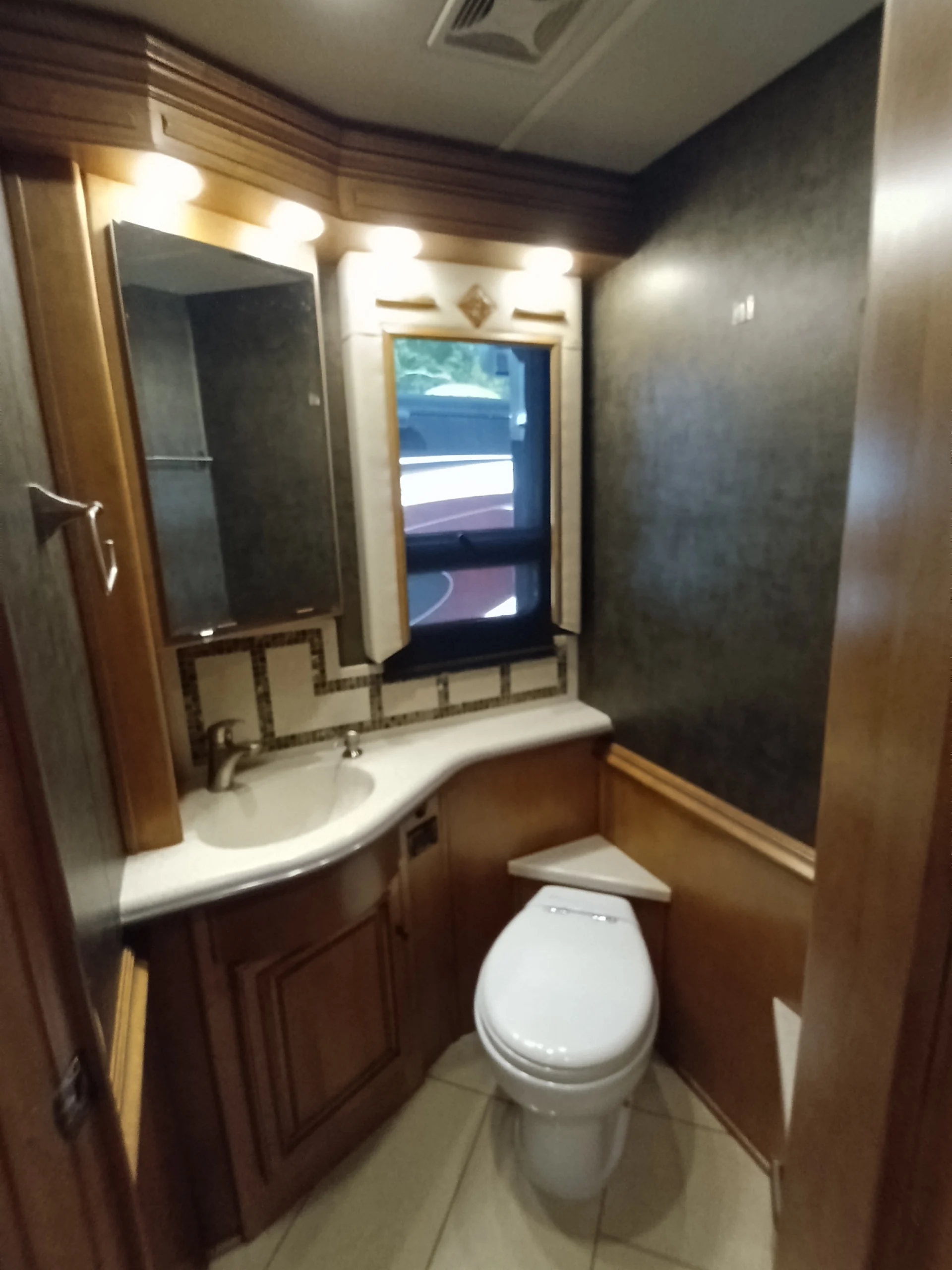 IMG_20260710_172722_269 Elegant motorcoach and RV bathroom with wooden cabinetry, a countertop sink, a toilet, and underlit mirror; window and tiled floor.