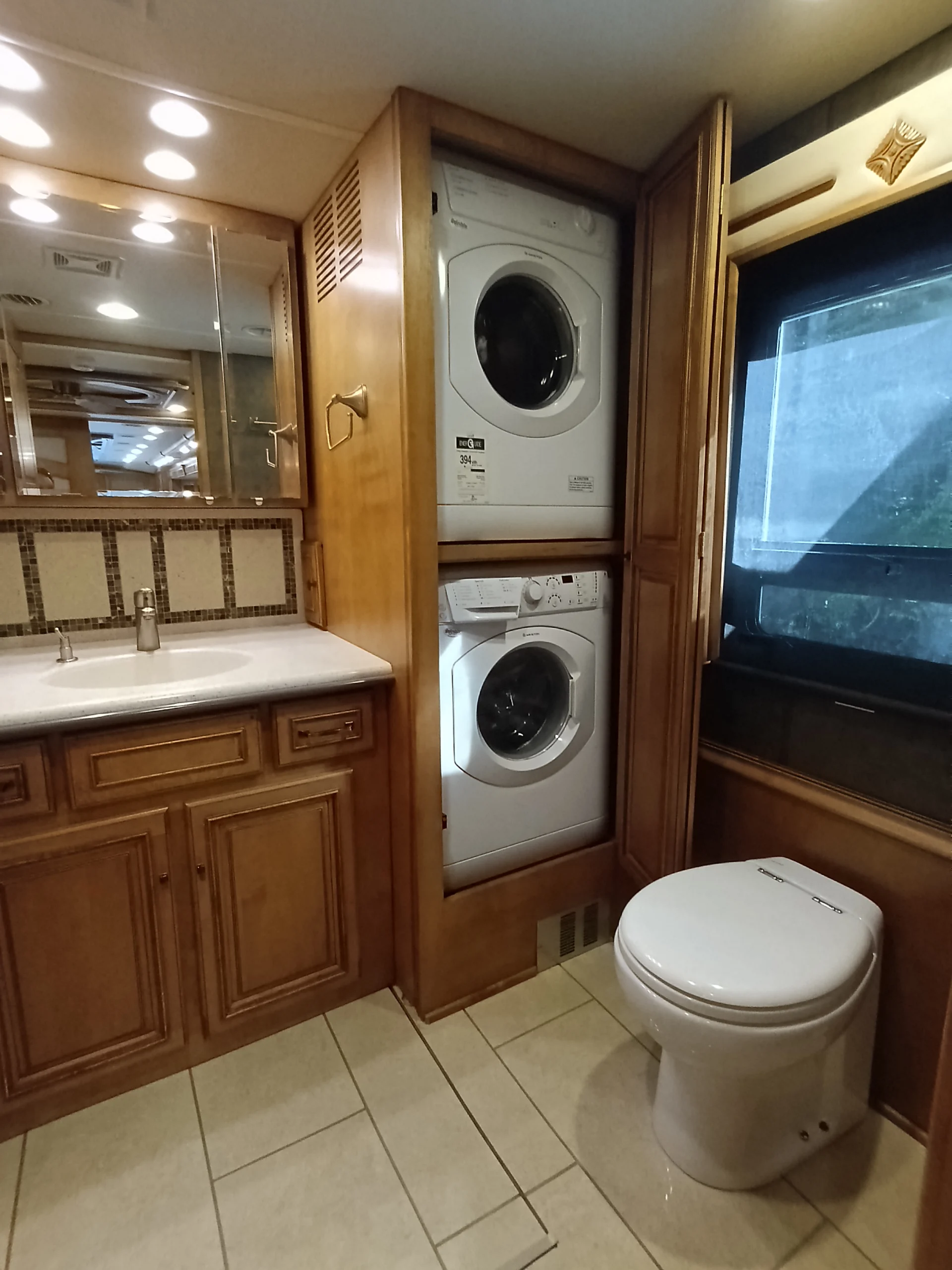 IMG_20260710_172903_307 RV bathroom featuring a stacked washer-dryer unit, wooden cabinets, a sink, and toilet. Suitable for Motorcoach and RVs.
