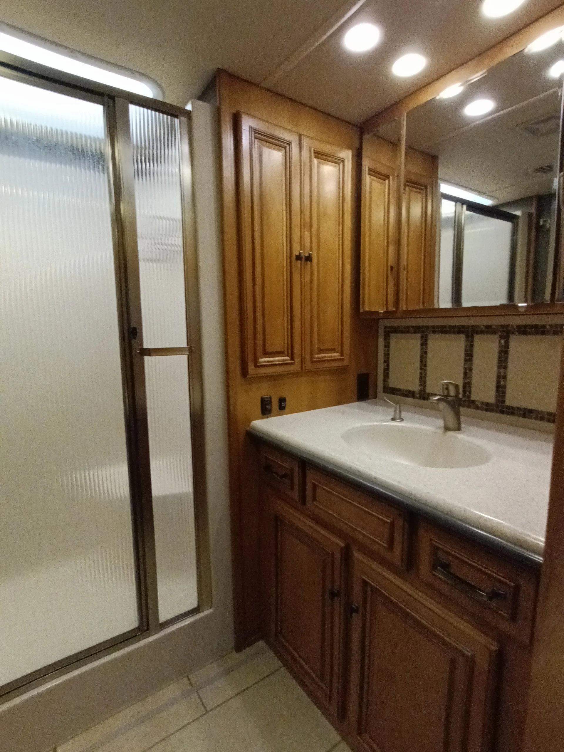 IMG_20260710_172928_002 RV bathroom featuring wood cabinets, large mirror, sink with beige countertop, and a shower with frosted glass door. Perfect for Motorcoach living.