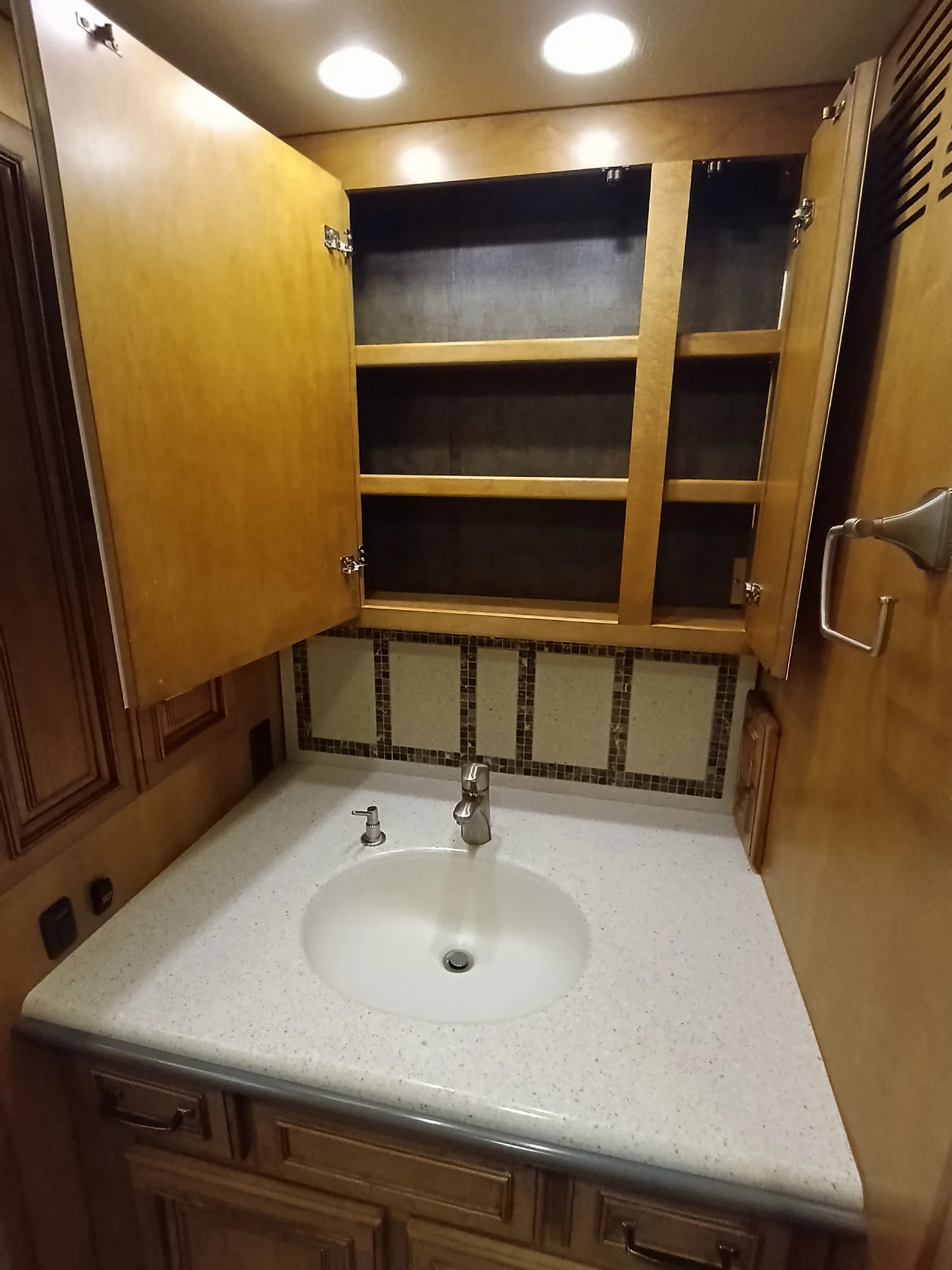 IMG_20260710_173007_878 RV bathroom vanity with open wooden cabinet, white sink, and mosaic tile backsplash. Ideal for Motorcoach interiors.