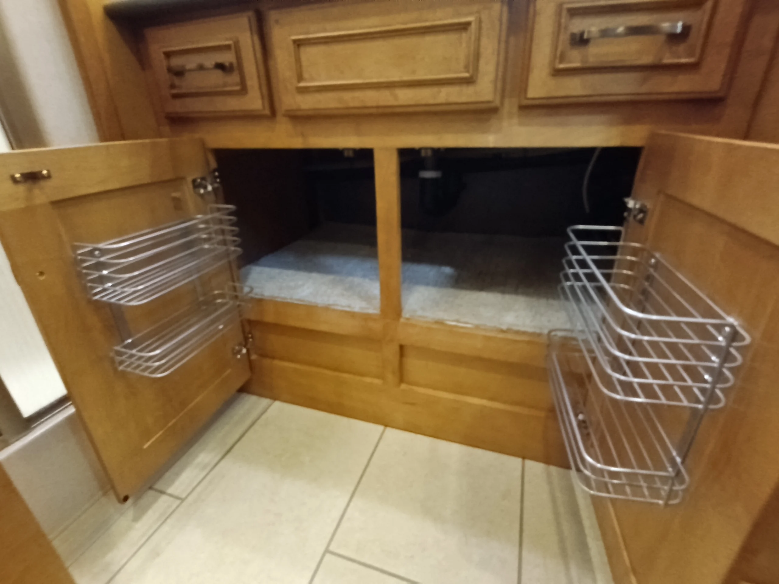 IMG_20260710_173145_730 Wooden cabinet with open doors featuring wire racks, typical storage in Motorcoach and RVs. White tiled floor underneath.