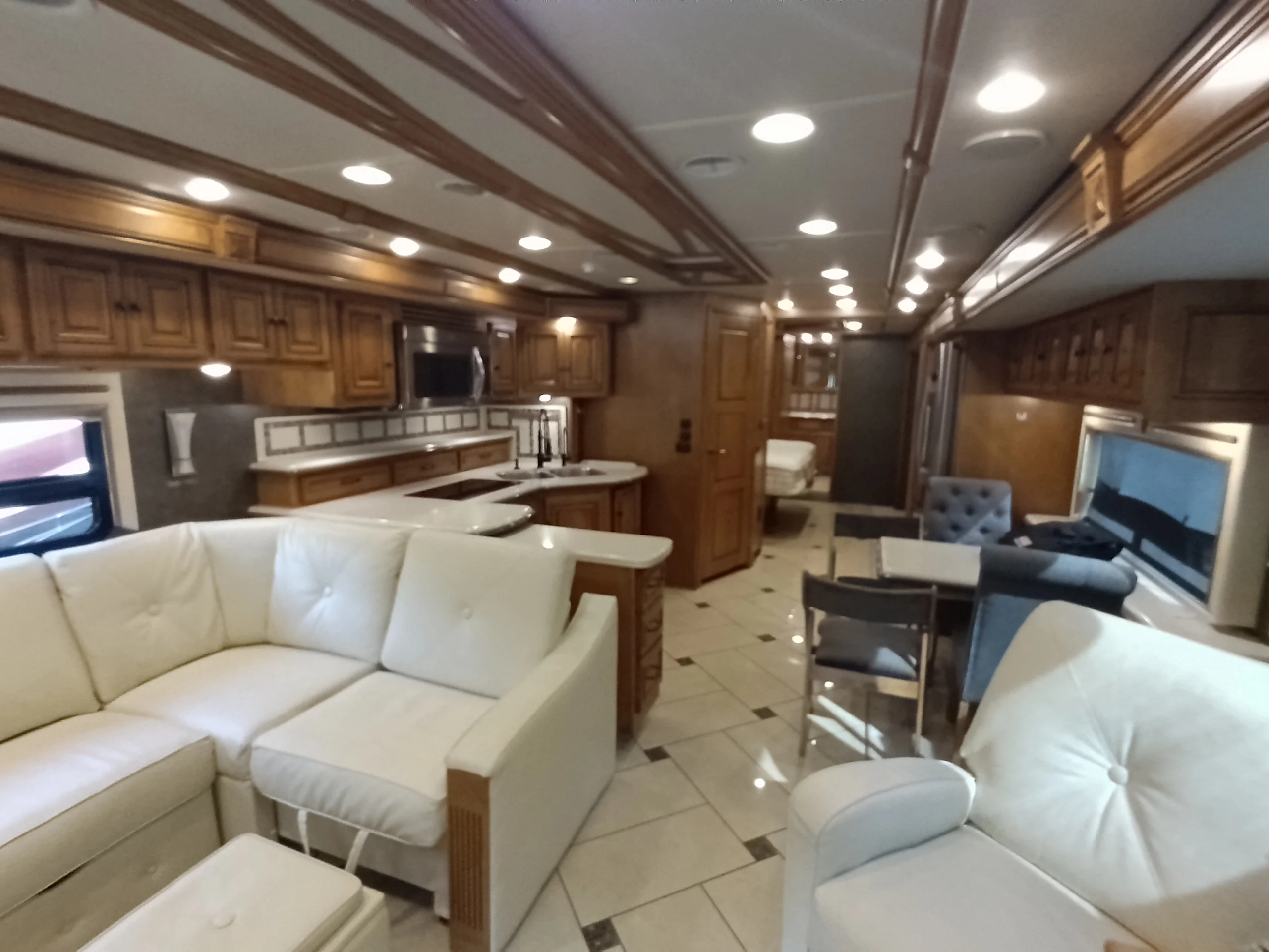 IMG_20260710_173231_538 Spacious Motorcoach interior with white seating, wooden cabinetry, dining area, and modern kitchen amenities.