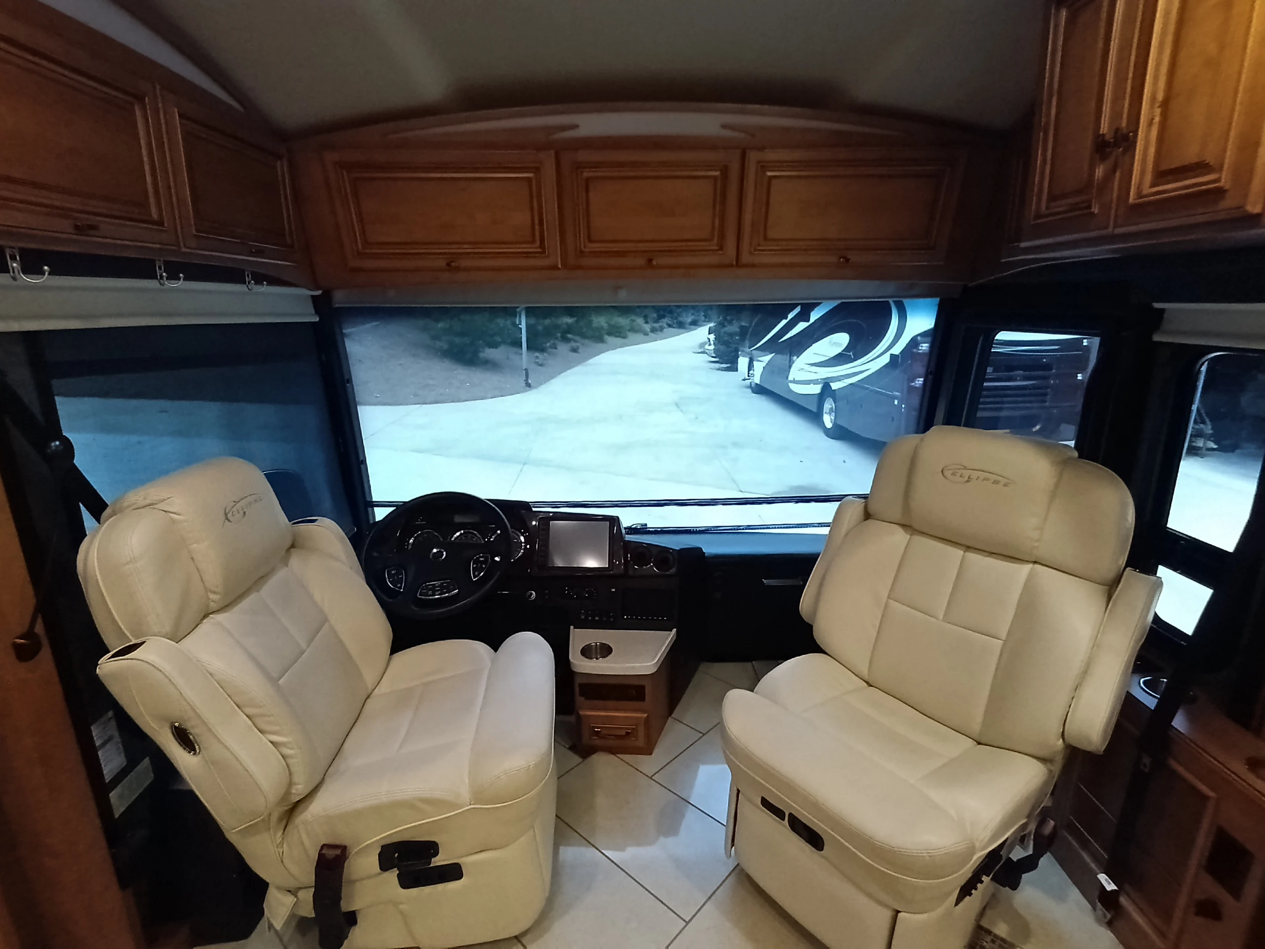 IMG_20260710_174845_240 Beige leather seats in a motorcoach's driver's area with large windshield view. Cabinets above and parked RVs visible outside.