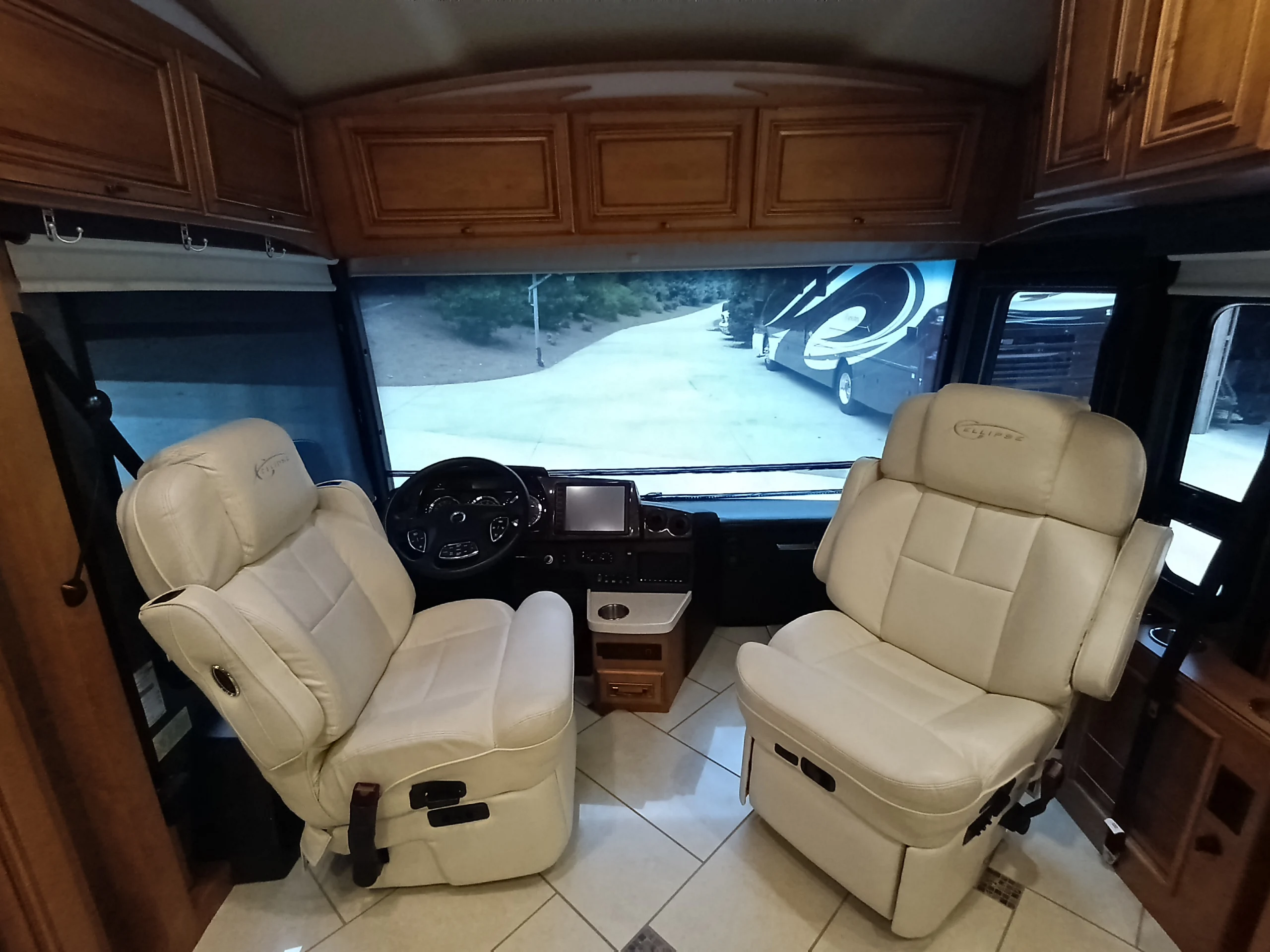 IMG_20260710_174851_640 Luxurious Motorcoach interior with two plush cream driver and passenger seats, panoramic windshield view, wooden cabinetry, and paved lot outside.