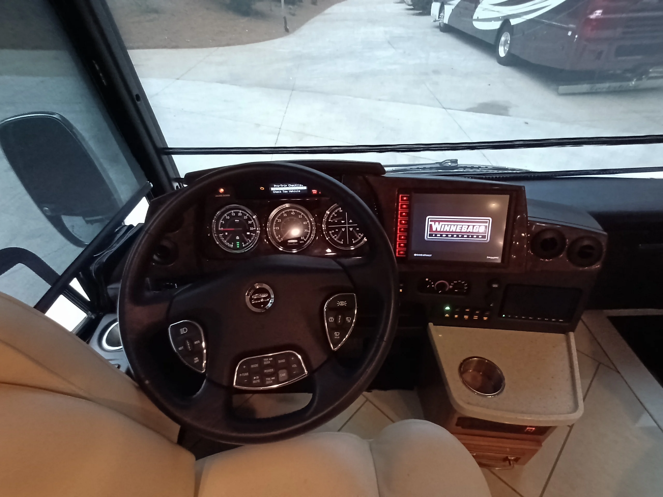 IMG_20260710_174916_112 Driver's seat and dashboard of a Motorcoach with steering wheel, gauges, electronic display, and cup holder, showing outside view through the windshie