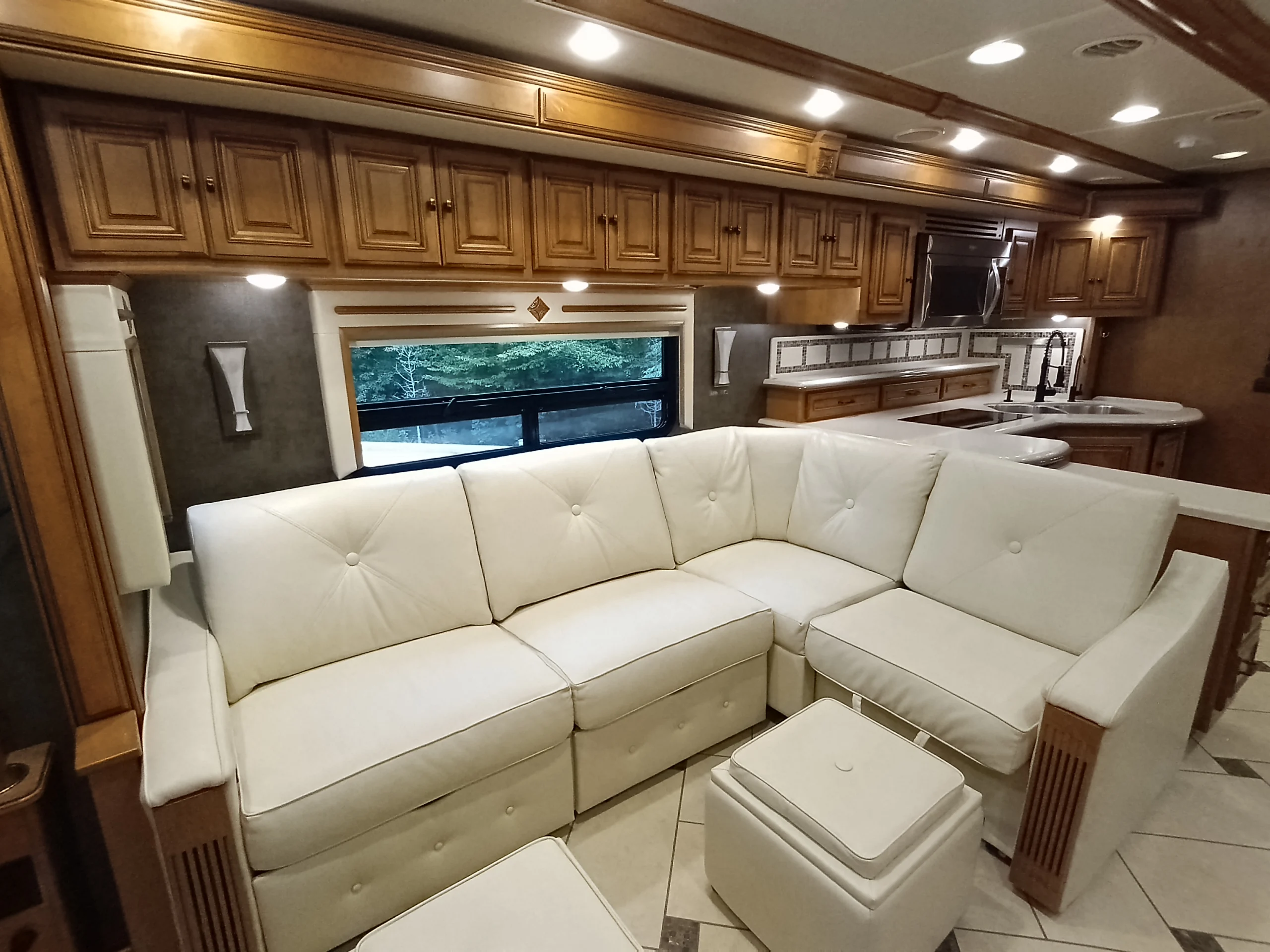 IMG_20260710_175058_919 Spacious Motorcoach interior with white leather sectional, wood cabinets, and stainless kitchen appliances.