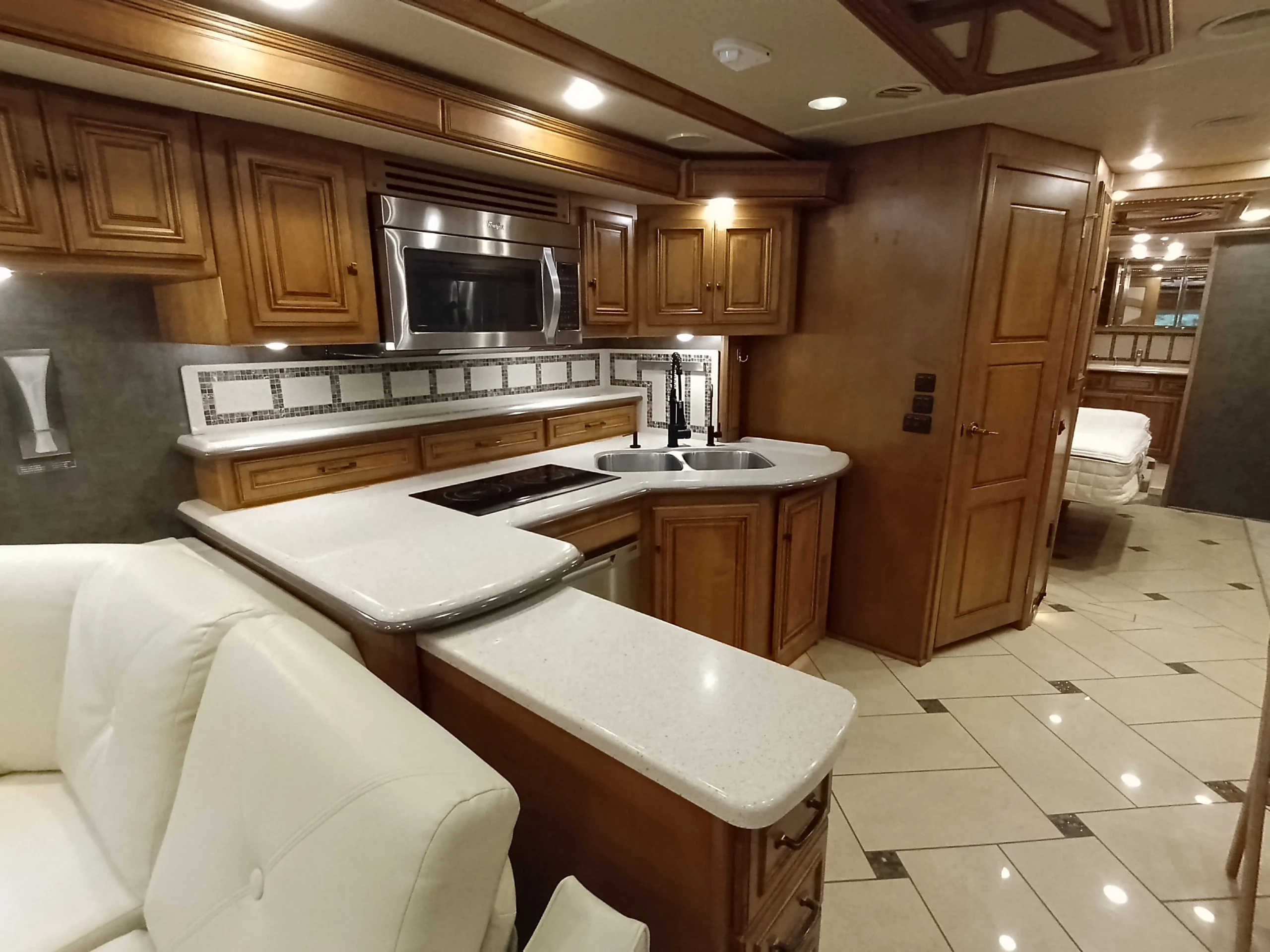 IMG_20260710_175105_804 Luxurious motorcoach interior with sleek kitchen, white countertops, double sink, and wooden cabinetry. Cream leather sofa in foreground.